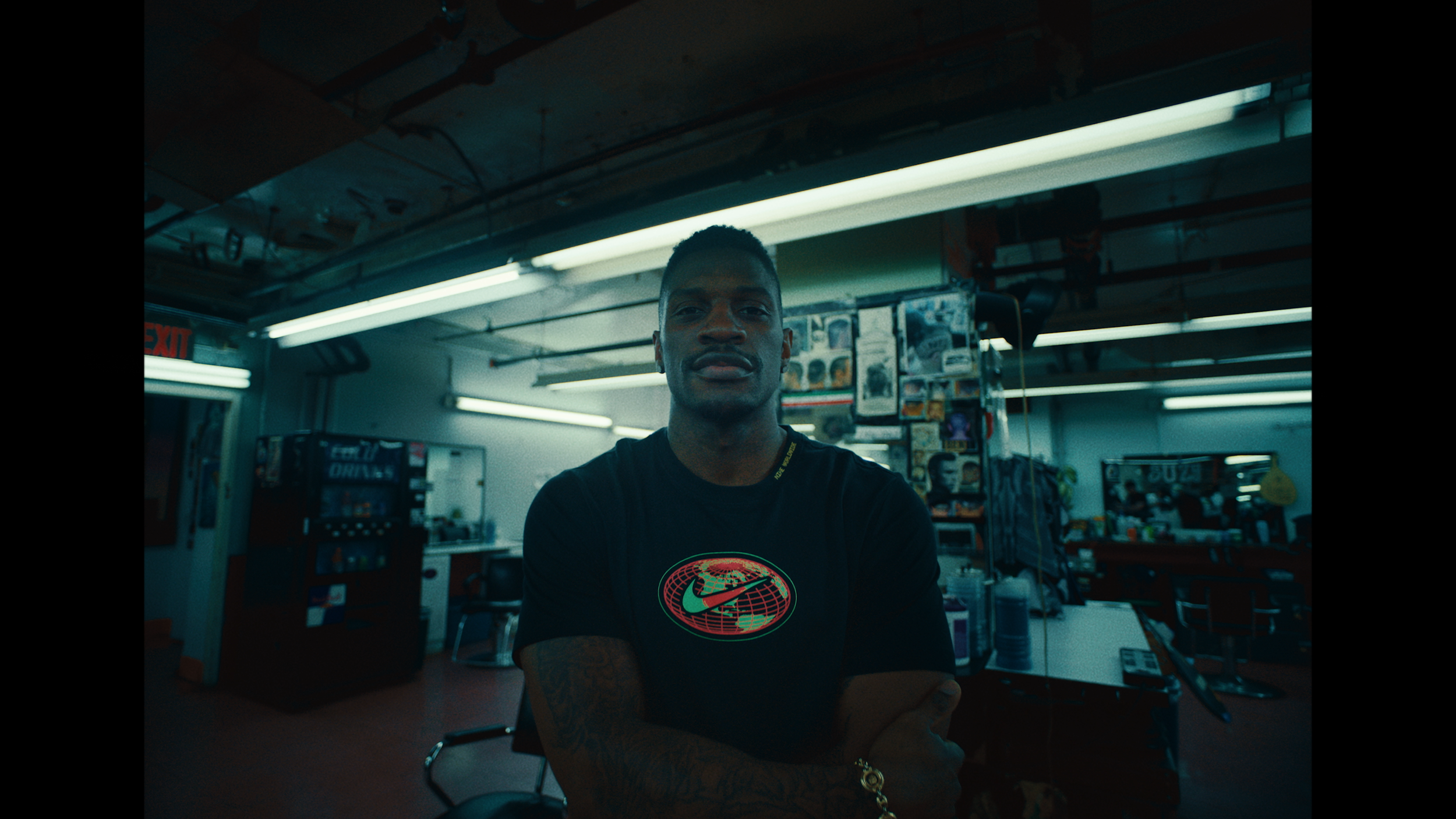 A man with tattoos on his arm stands with arms crossed in a tattoo shop, illuminated by fluorescent lights, with tattoos and posters on the walls behind him.