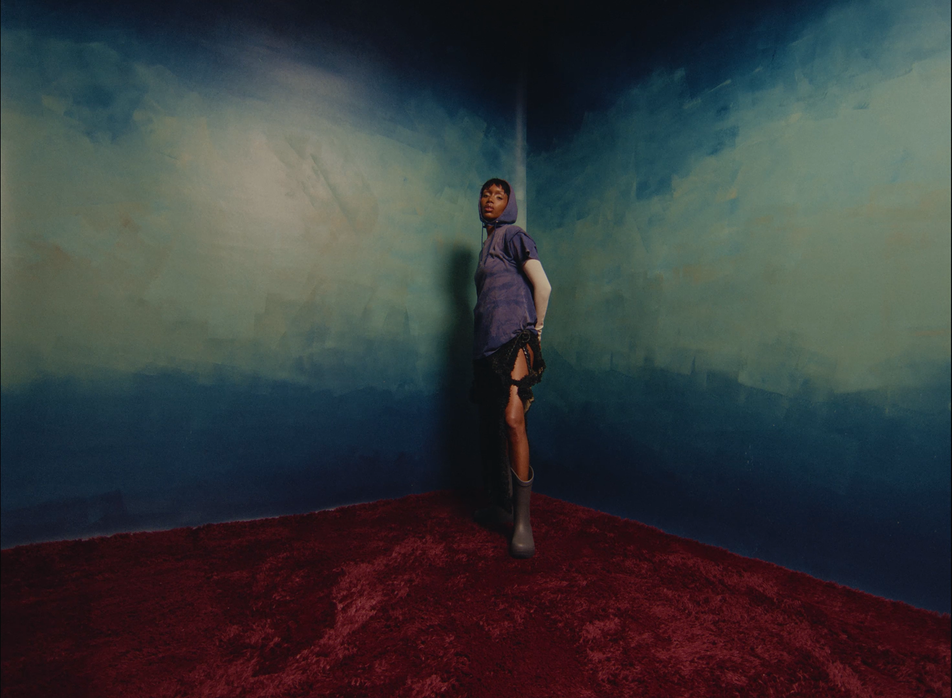 A woman standing in the corner of a room with blue and green wall art, wearing a purple hooded shirt, black skirt, and gray rain boots on a red carpet.