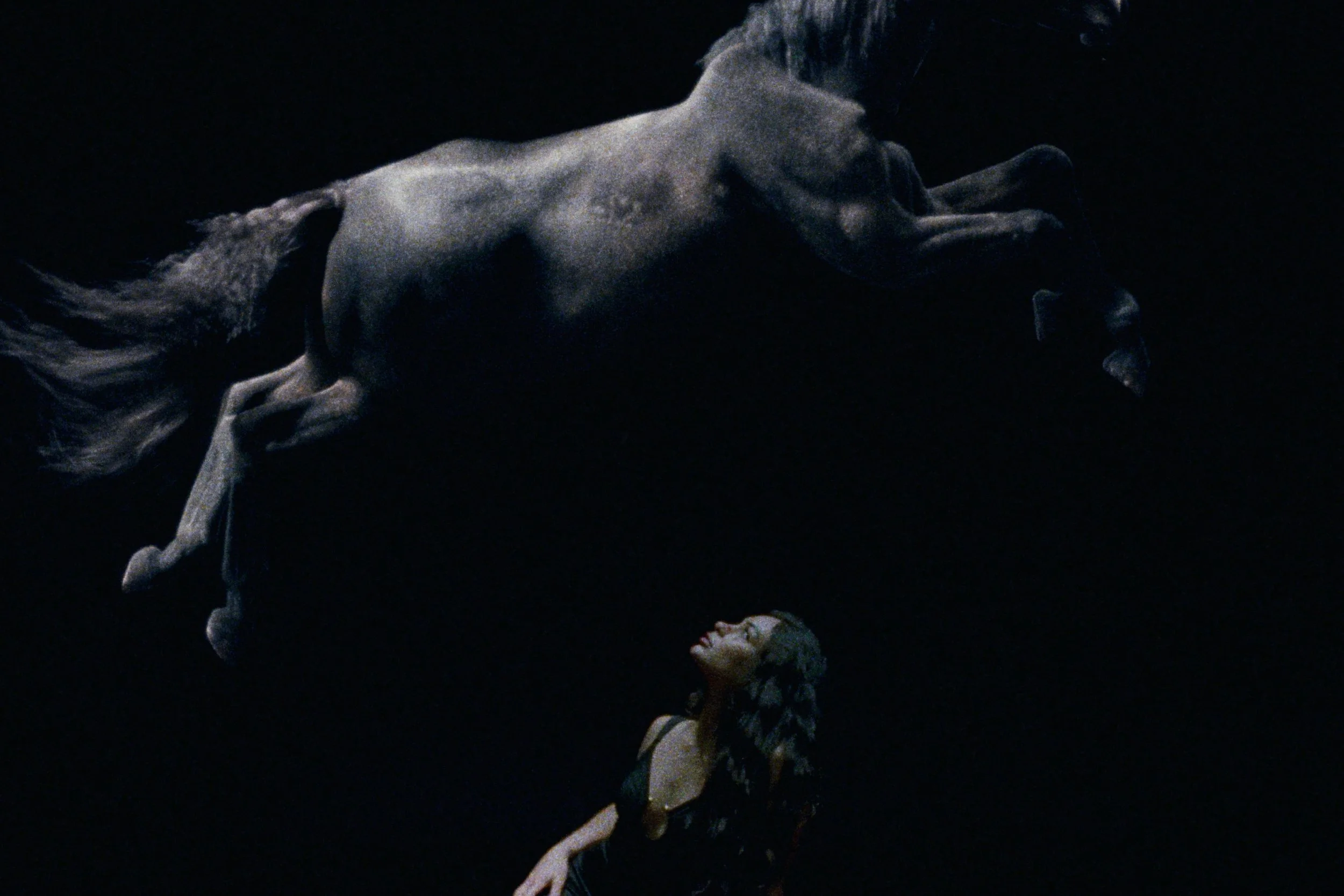 A woman in a black dress looking up at a dark gray horse floating above her in a black background.