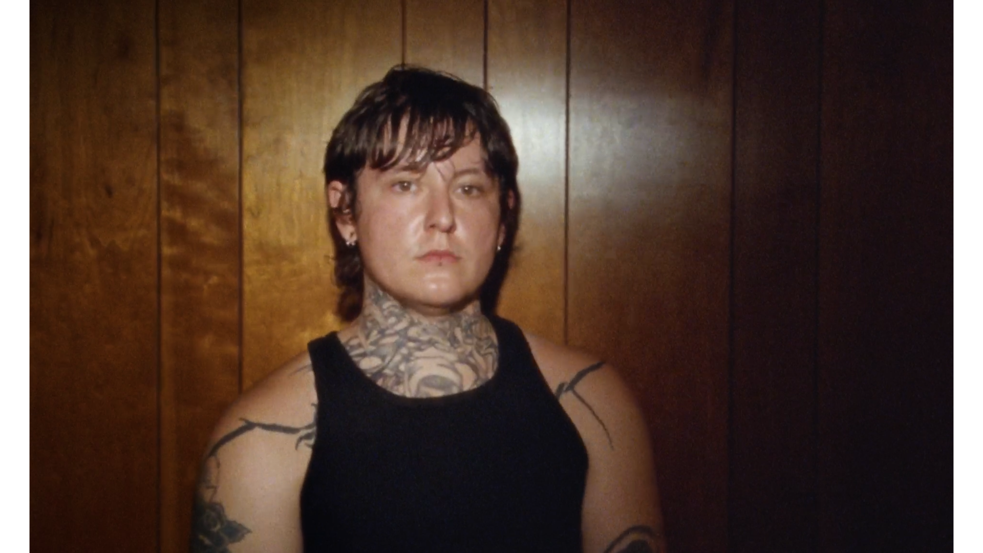 A person with short dark hair, tattoos on their neck and arms, wearing a black sleeveless shirt, standing against a wooden panel wall.