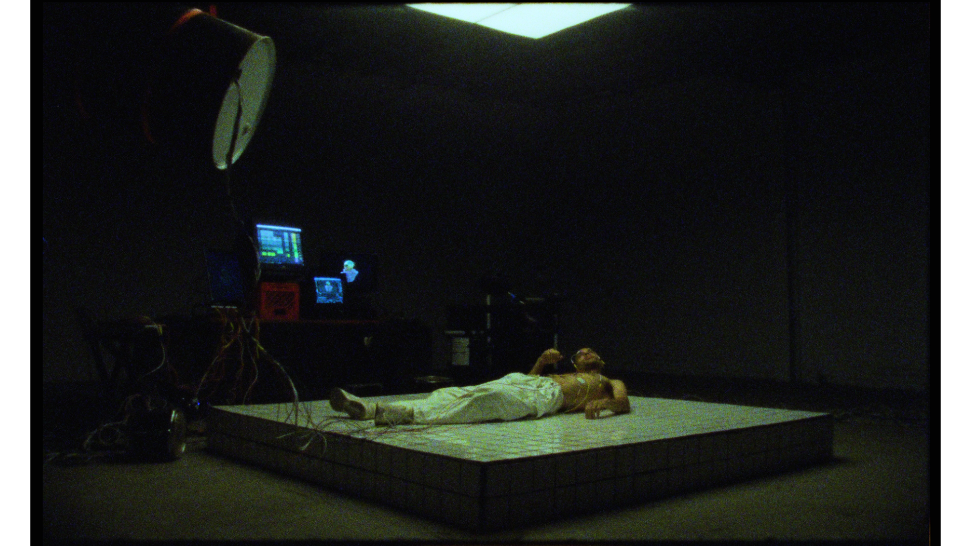 A person wearing white pants and a yellow top lying on a raised platform, with monitors and equipment in a dark room.