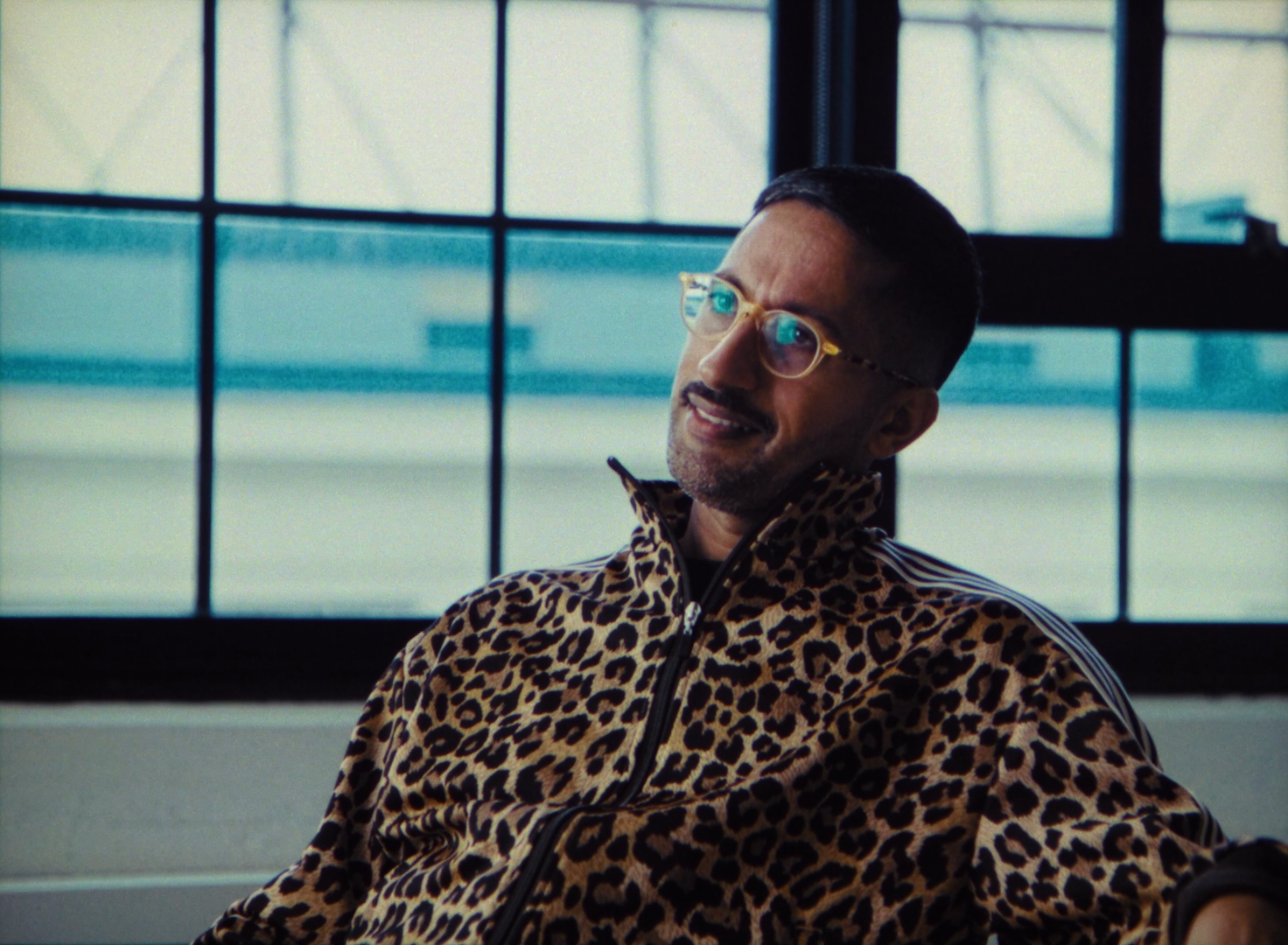 A man wearing glasses and a leopard print jacket sitting by a large window, looking slightly to the side and smiling.