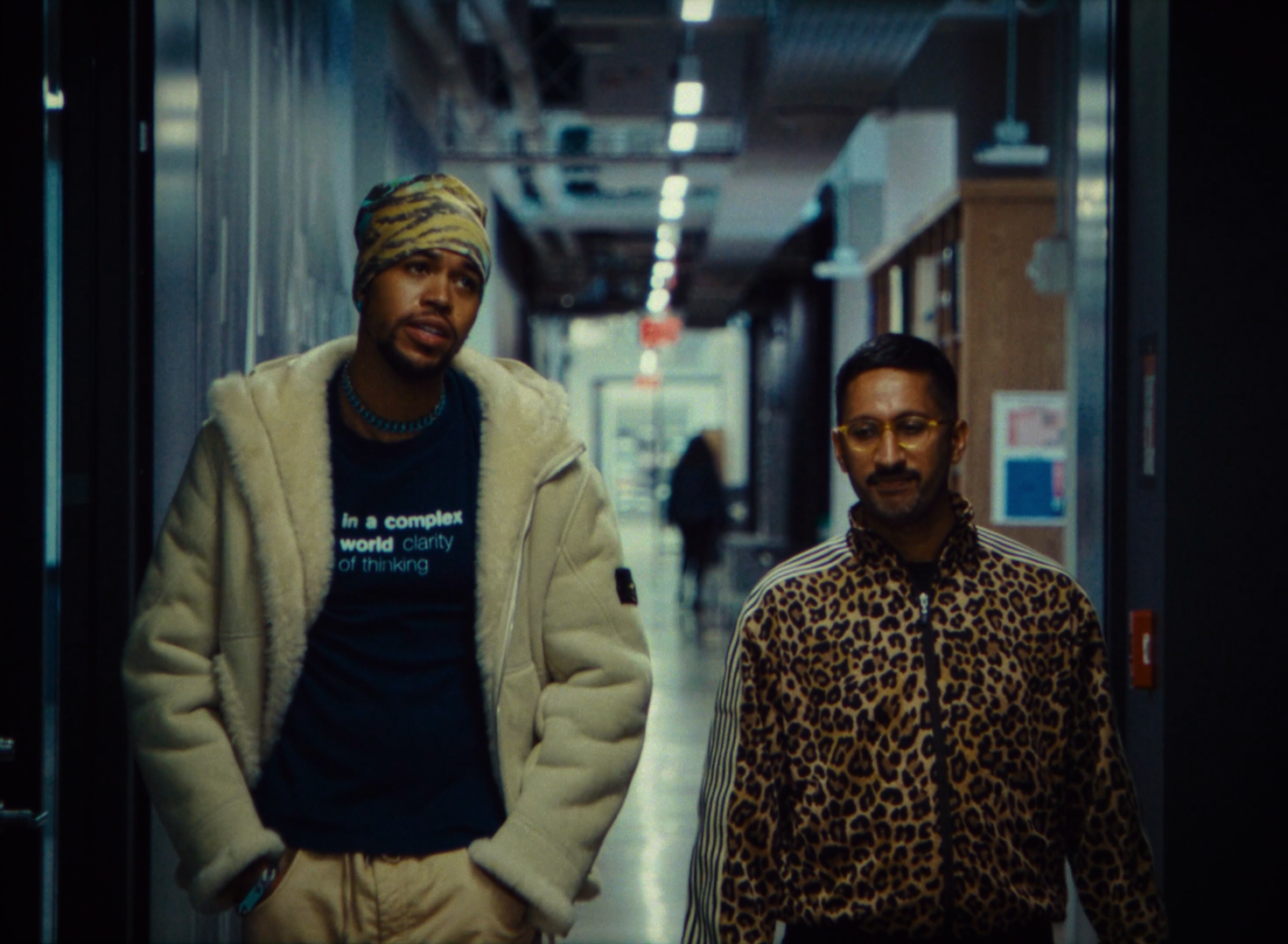 Two men walking down a hallway inside a building, one wearing a beige jacket and a headscarf, and the other in a leopard print shirt and glasses.