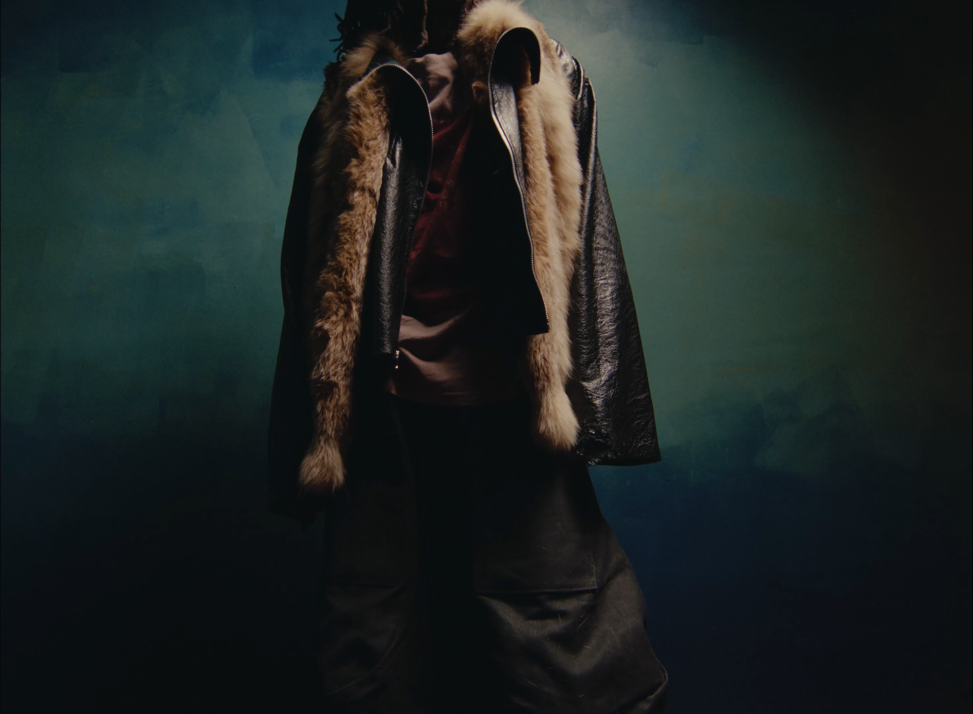 A person wearing a black leather jacket with a fur lining, a brown shirt, and dark pants, standing against a dark, gradient background.