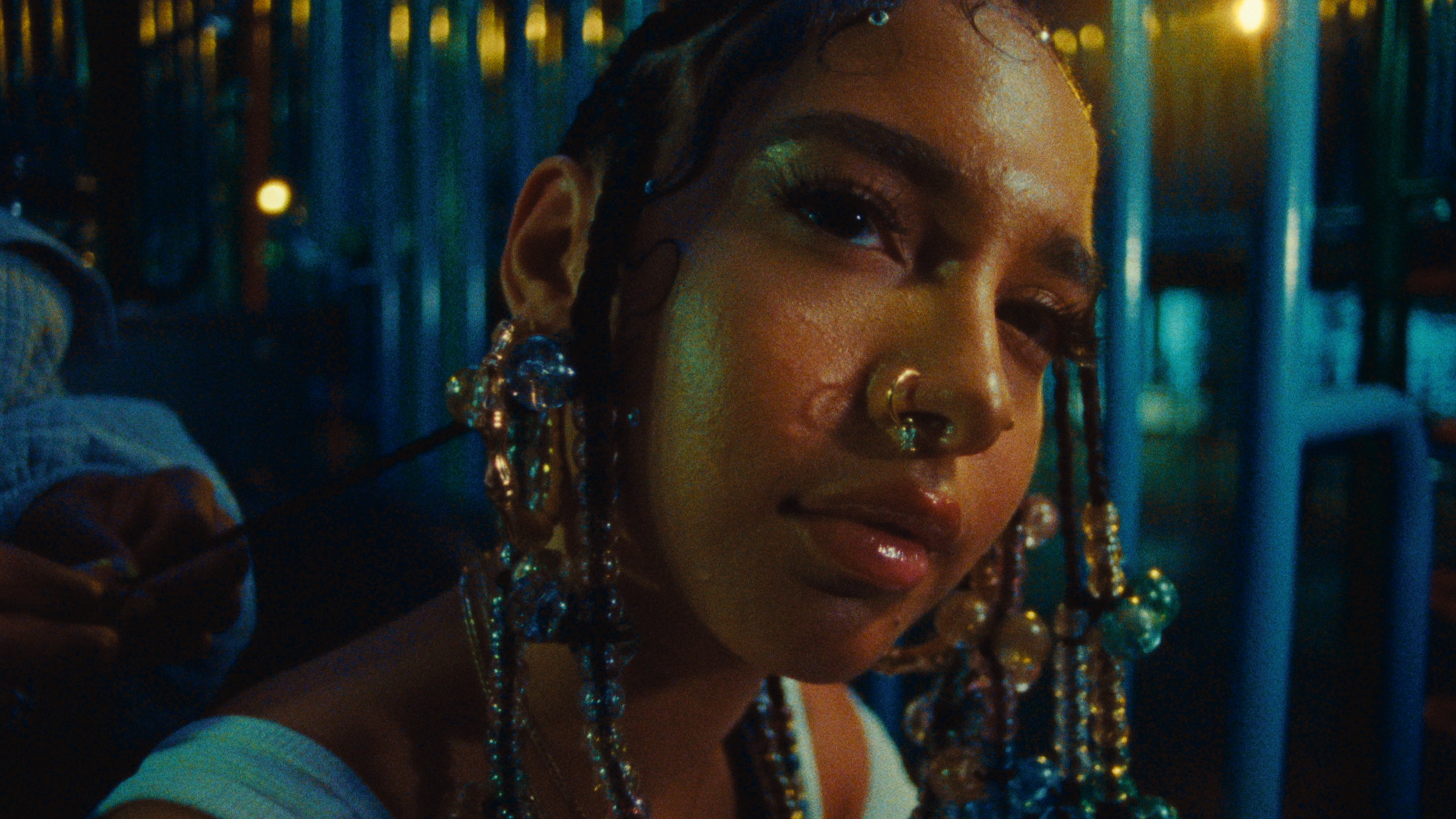 A young woman with braided hair adorned with beads and jewelry, wearing hoop earrings and a nose ring, looking at the camera with a confident expression in a dimly lit setting with blue and yellow lights.