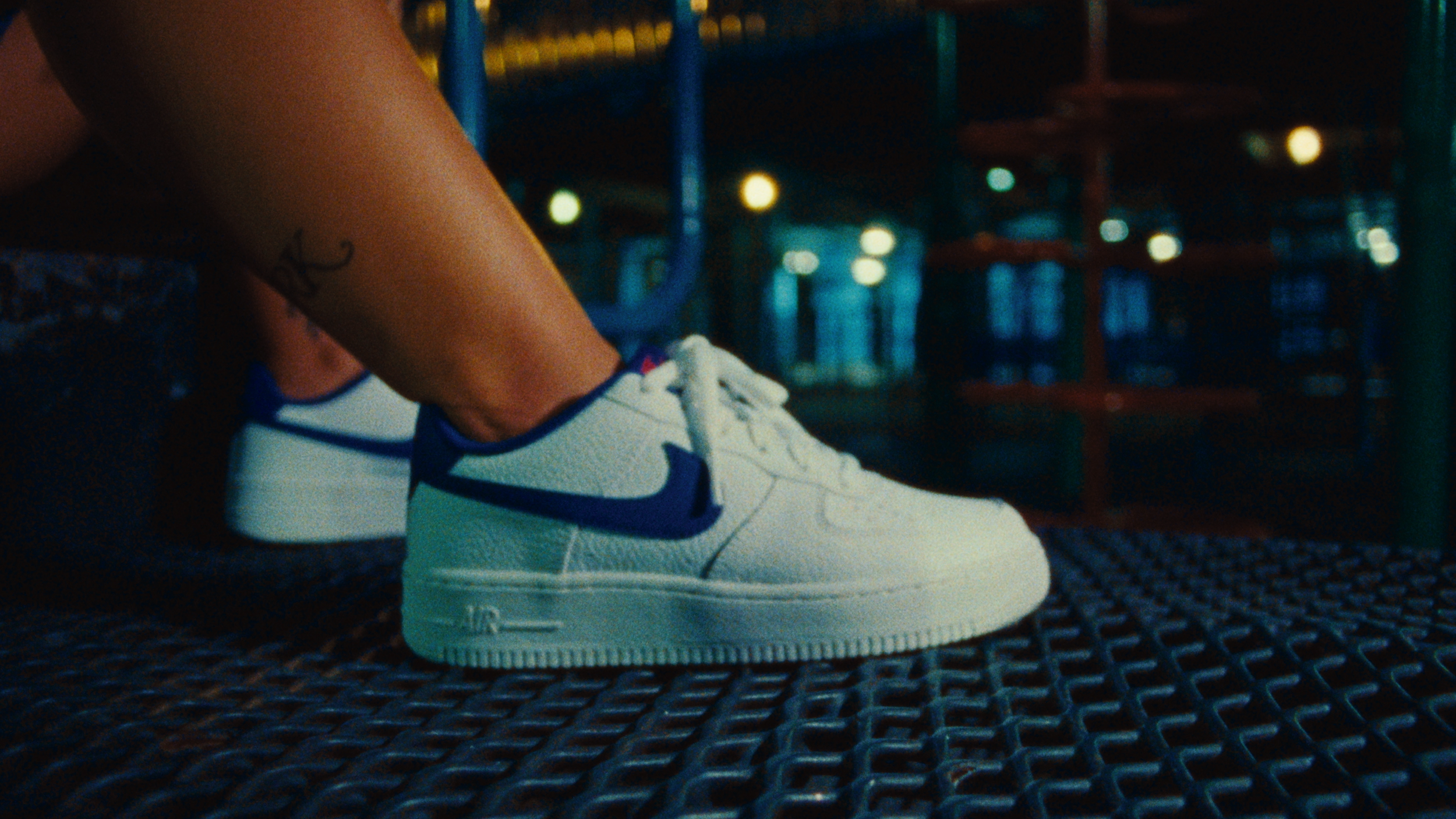 Close-up of a person's foot wearing a white Nike sneaker with purple accents, on a metal grate at night with blurred lights and playground equipment in the background.