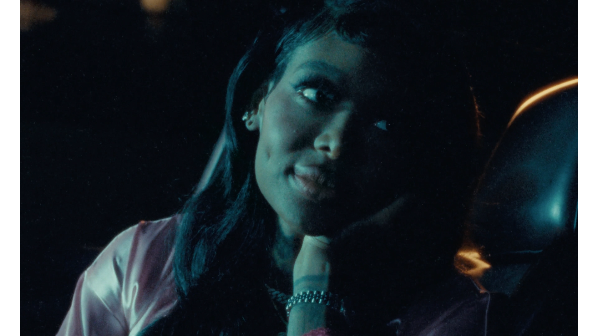 A close-up of a woman's face in a dimly lit environment, with her head slightly tilted and covering part of her face with her hand, wearing a pink satin jacket and a wristwatch, looking at the camera.