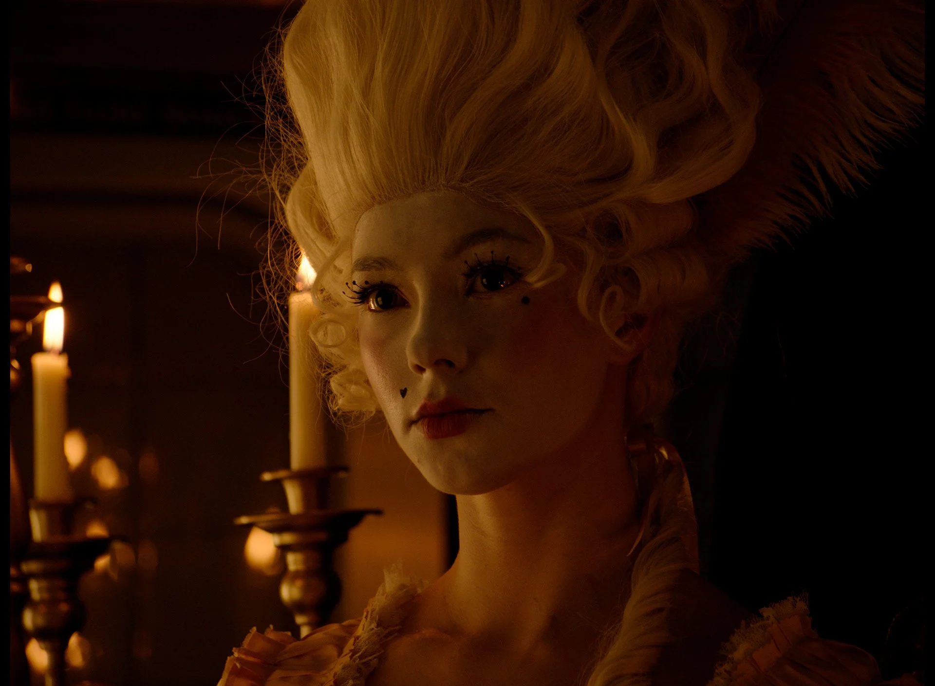 A woman with elaborate, curly blonde hair, makeup including long eyelashes, a black heart mole on her cheek, and wearing a high-collared Victorian-style dress. She is in a dimly lit room with candles in the background.