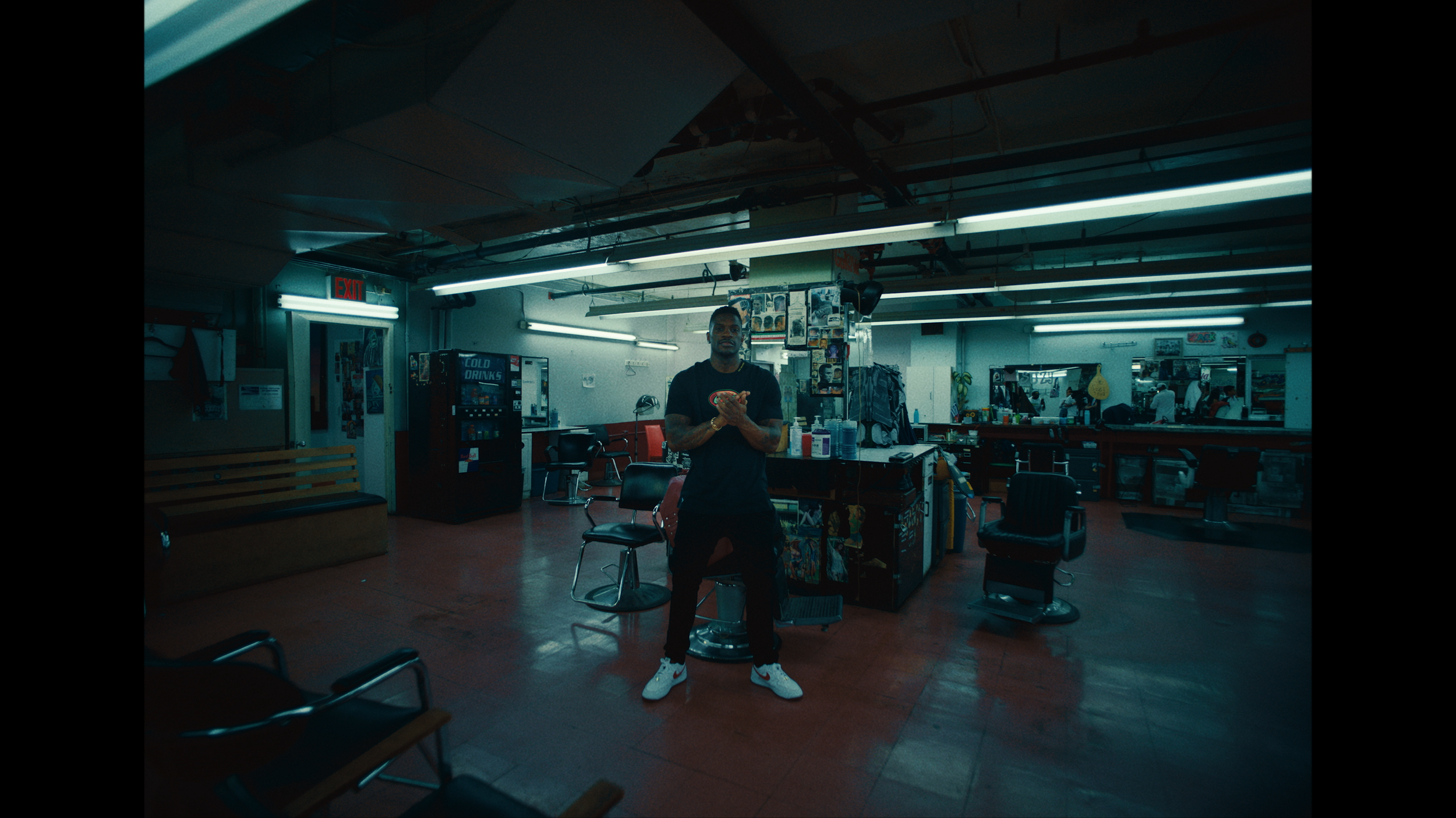 A man standing in a barbershop, holding a phone, with barber chairs, mirrors, and various salon tools in the background.