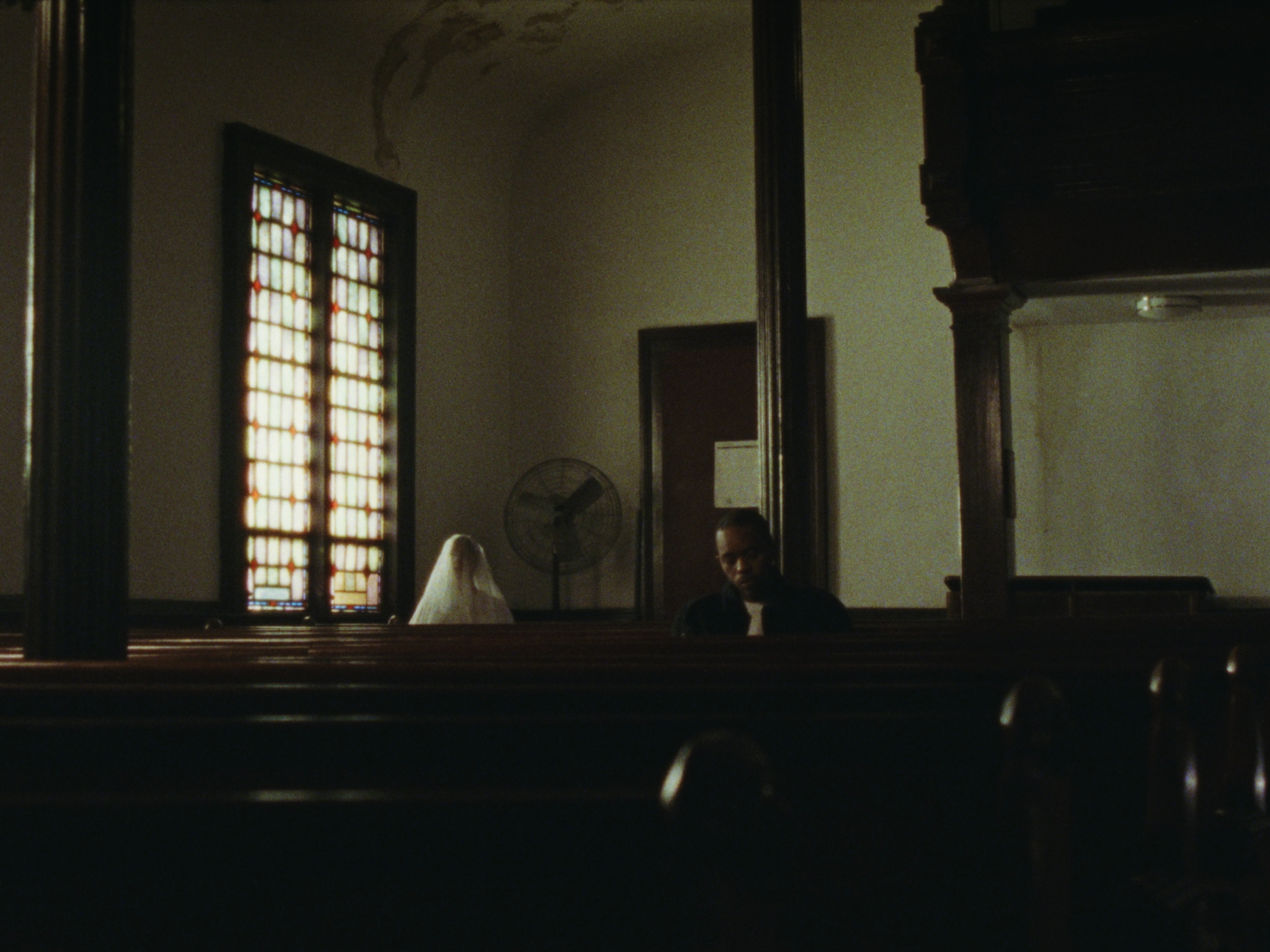 A man sitting in a church pew, with a ghost-like figure in white behind him near a church window with stained glass.