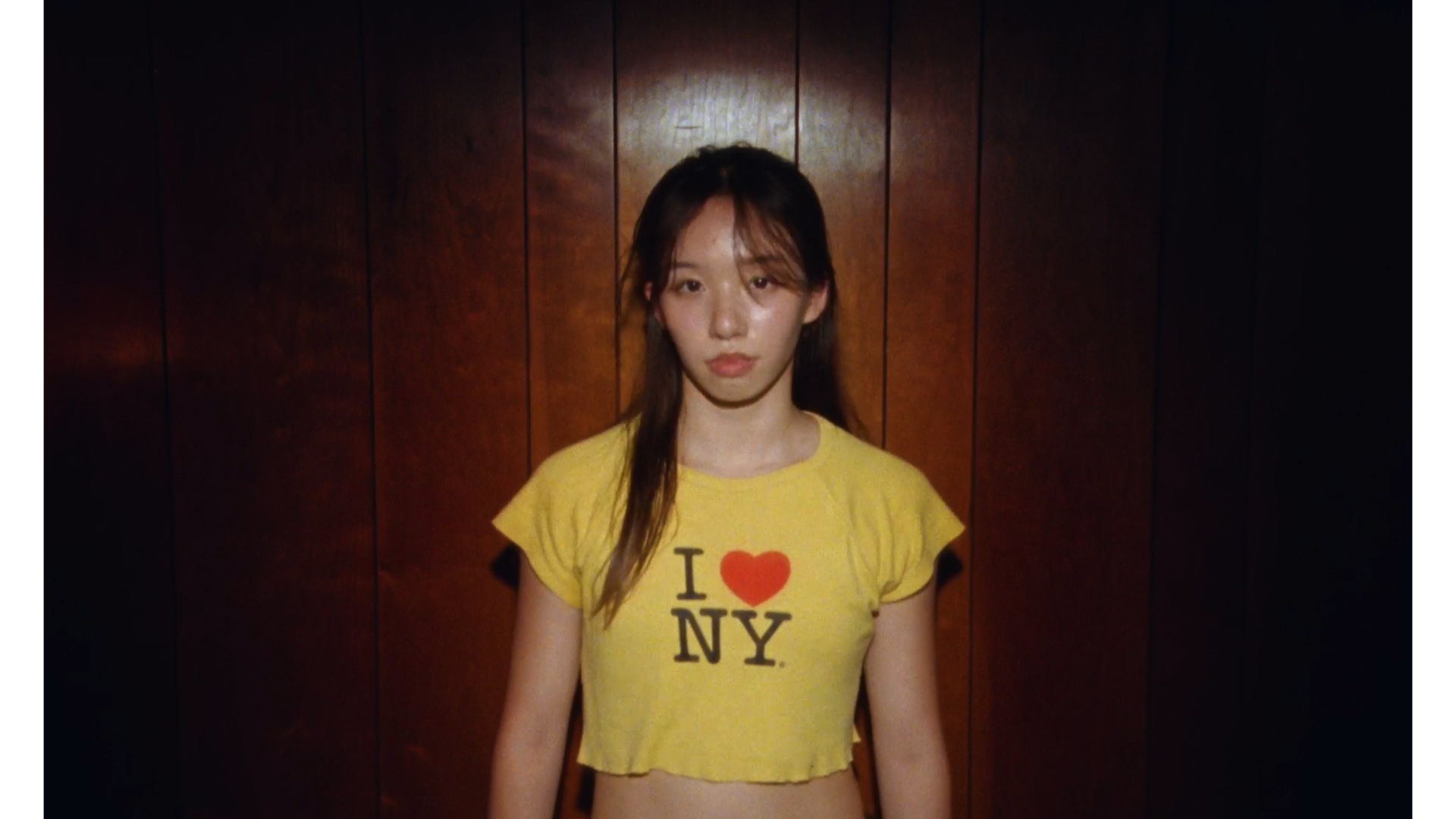 A young woman with long hair standing against a wooden wall, wearing a yellow T-shirt that says 'I heart NY' and looking directly at the camera.