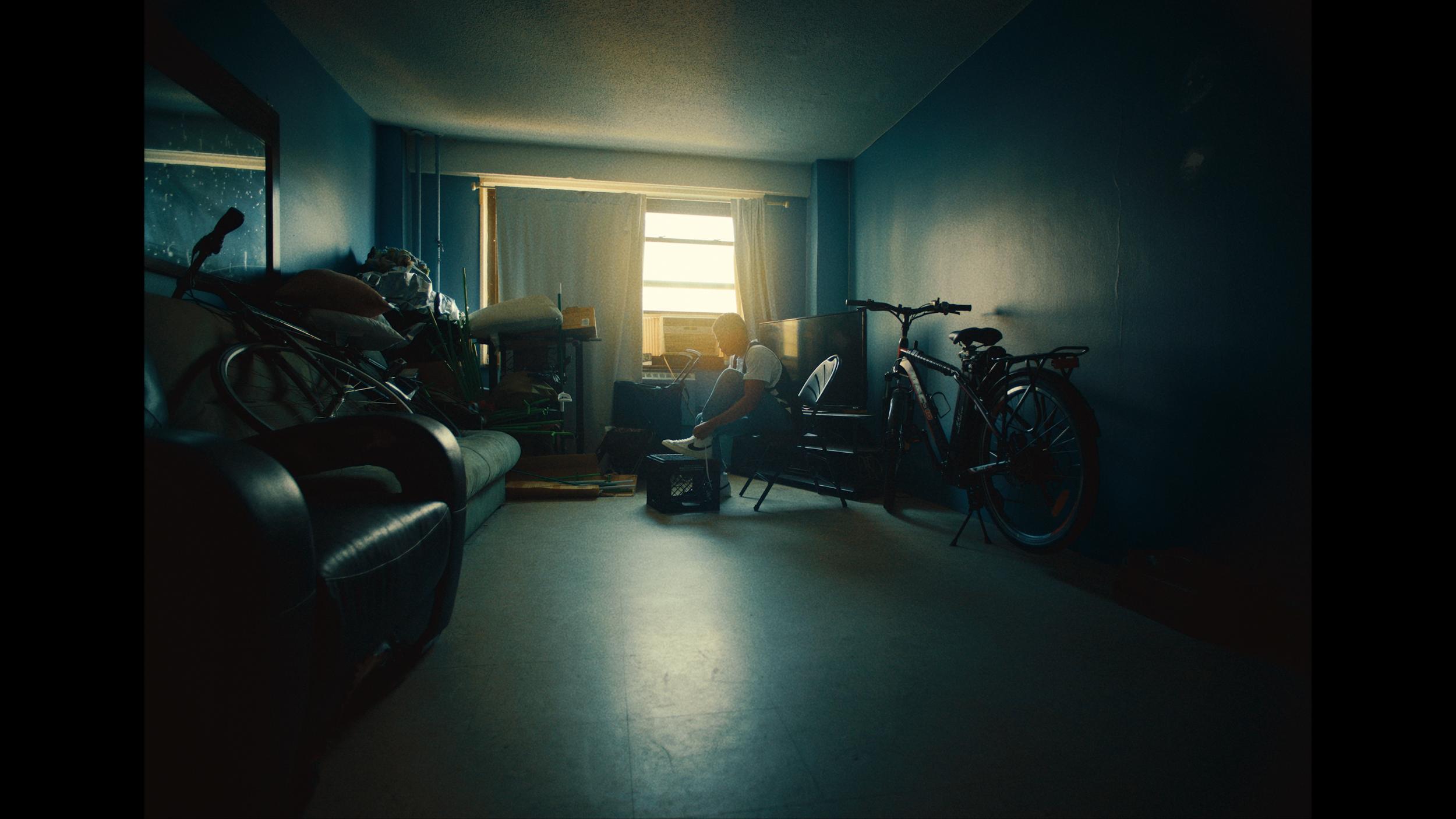 A person sits on a chair near a window in a dimly lit room, surrounded by a bicycle, a sofa, and various clutter, with sunlight coming through the window.