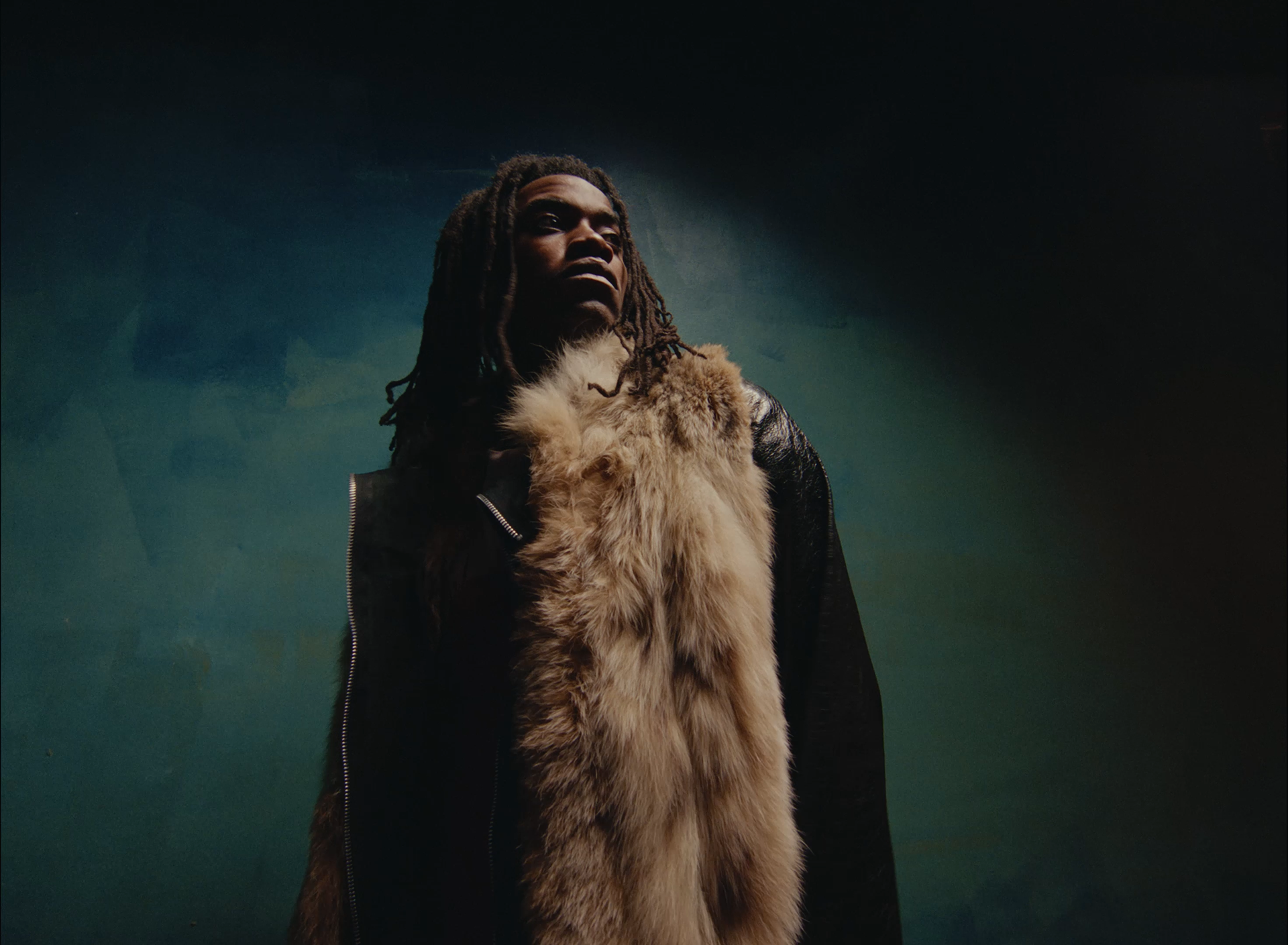 A woman with dreadlocks looking upwards, wearing a black jacket and a large fur stole against a dark, gradient background.