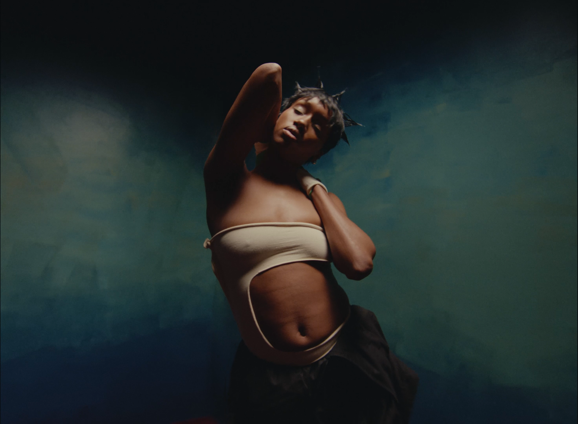 Person with dreadlocks posing with one arm behind head against a dark, textured background.