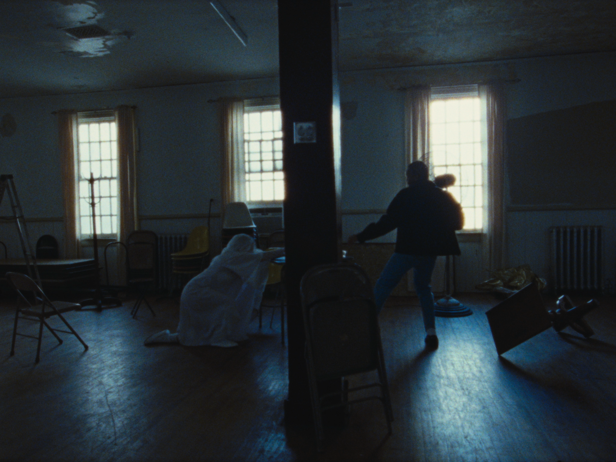 A dimly lit room with chairs and tables, two people are present with one sitting on the floor in a hoodie and the other standing near a window, possibly holding an object, with sunlight coming through the window.