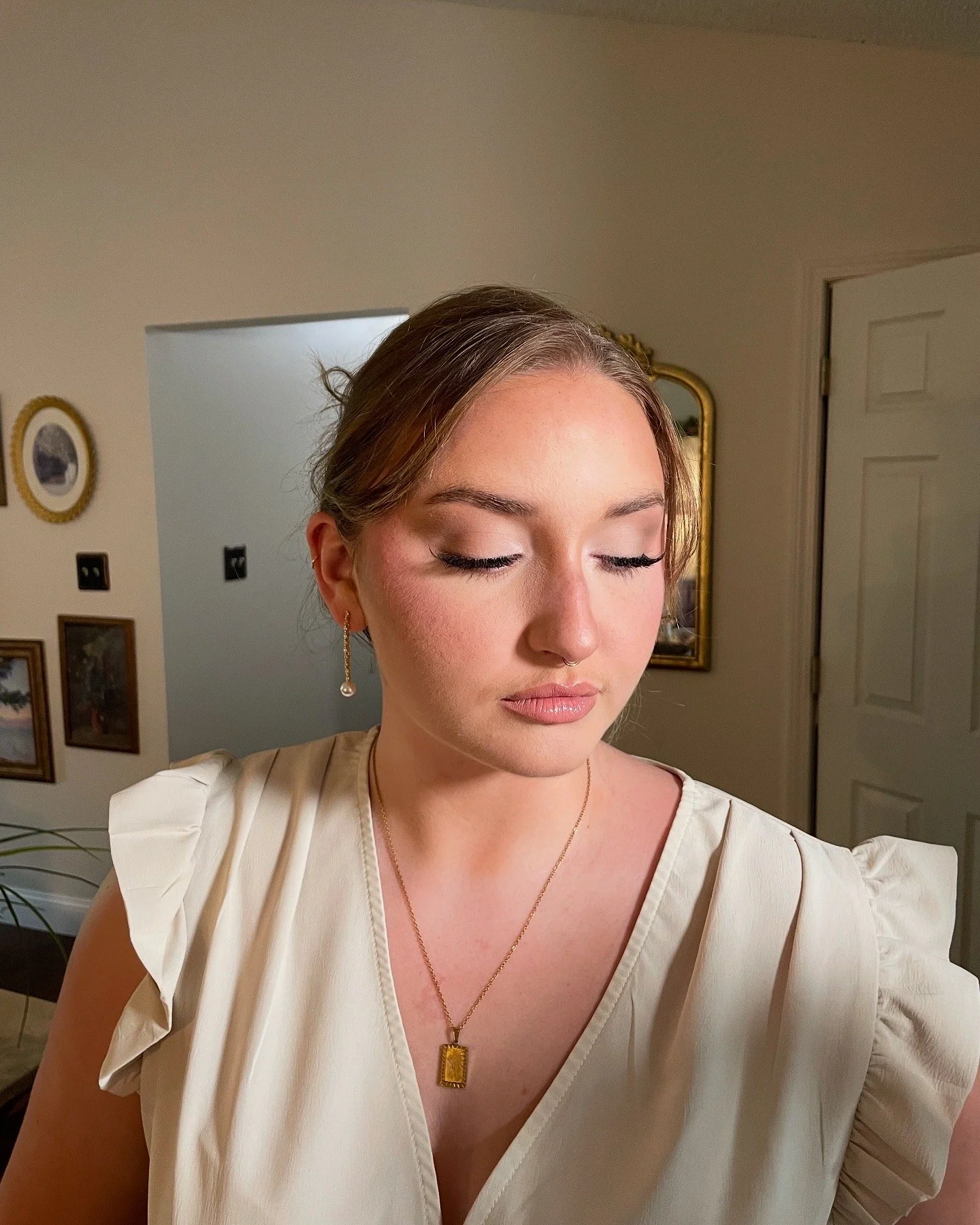 A woman with her eyes closed, wearing makeup, gold jewelry, and a cream-colored dress with ruffled sleeves, standing in a room with framed pictures and a mirror in the background.