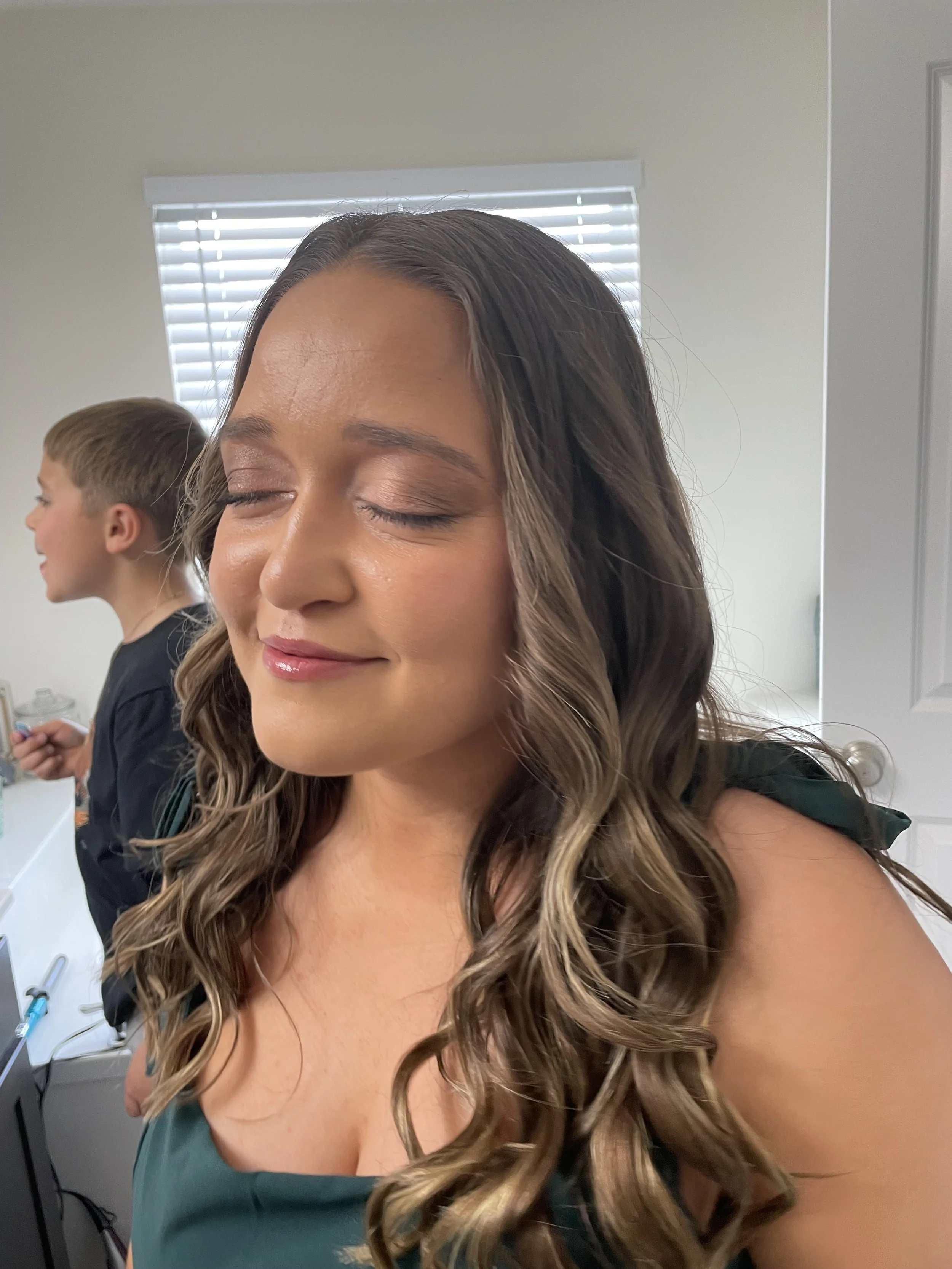 Woman with long wavy brown hair and closed eyes, smiling, in a room with a boy in the background near a bathroom mirror.