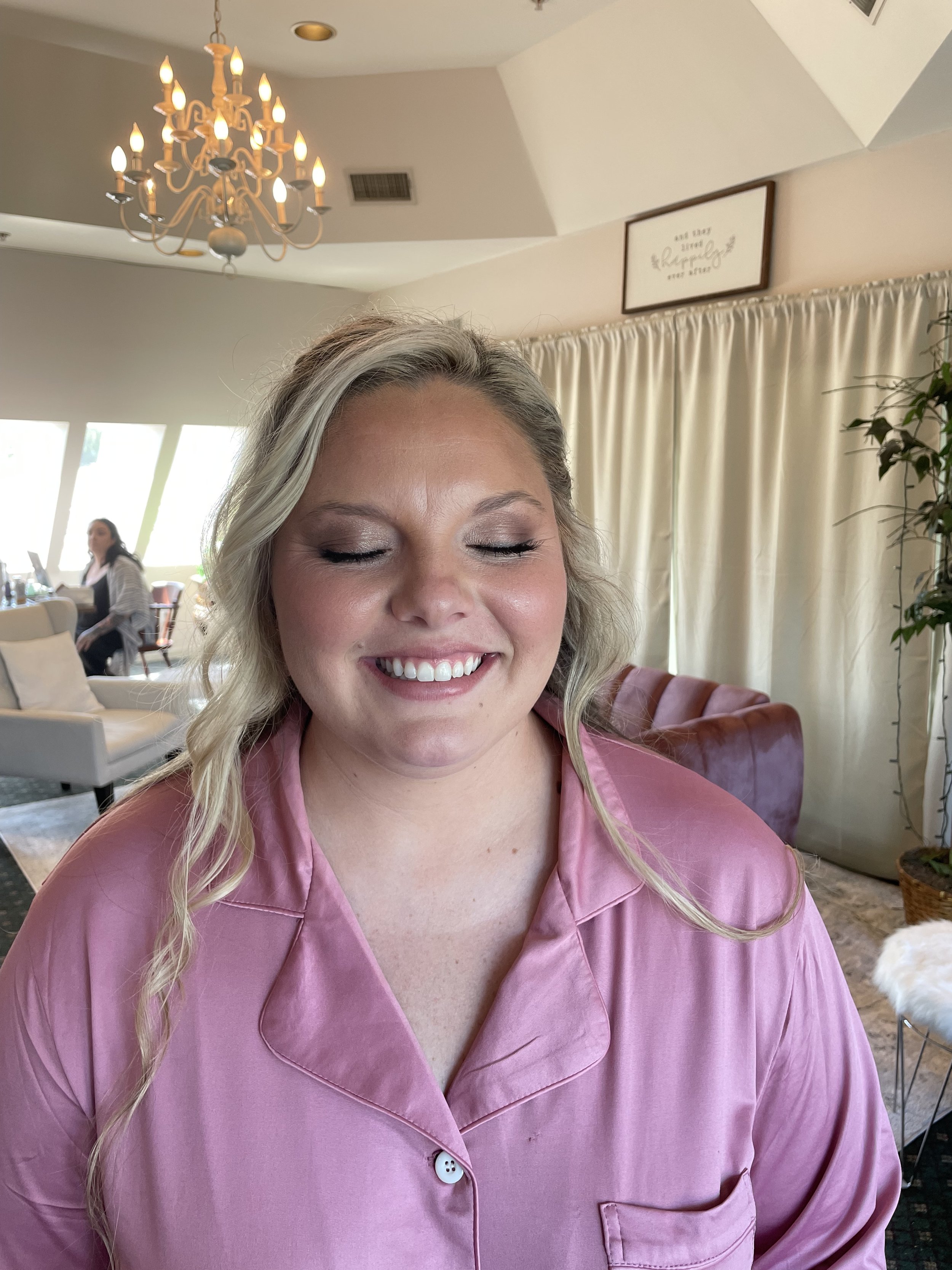 A woman with blonde hair and closed eyes smiling, wearing pink pajamas, in a well-lit room with a chandelier, beige curtains, and a mix of modern and vintage furniture.