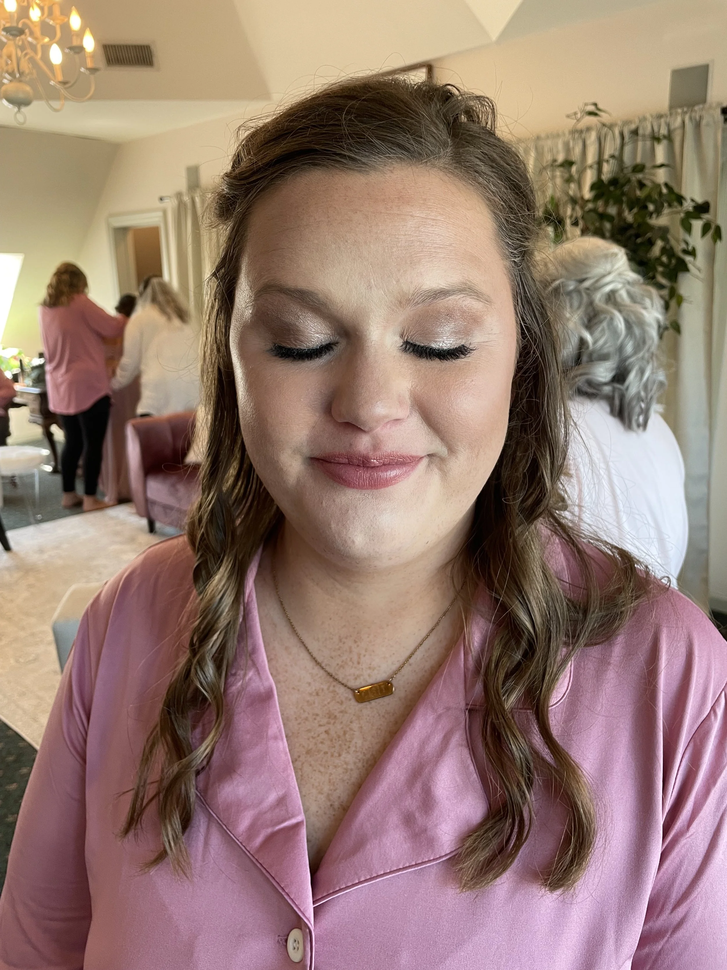 A woman with closed eyes and wavy brown hair in a pink satin robe, wearing a gold necklace, in a room with other women preparing and styling hair.