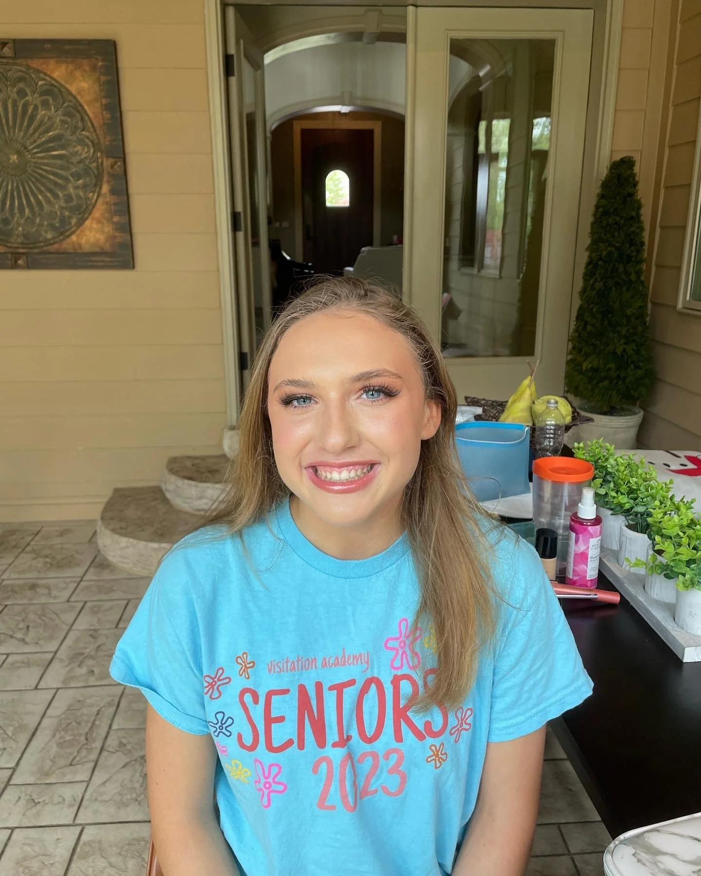 A young woman with blonde hair and blue eyes smiling, wearing a blue shirt with 'SENIORS 2023' and colorful decorations, sitting at an outdoor table on a porch with potted plants, spray bottles, and other items in the background.