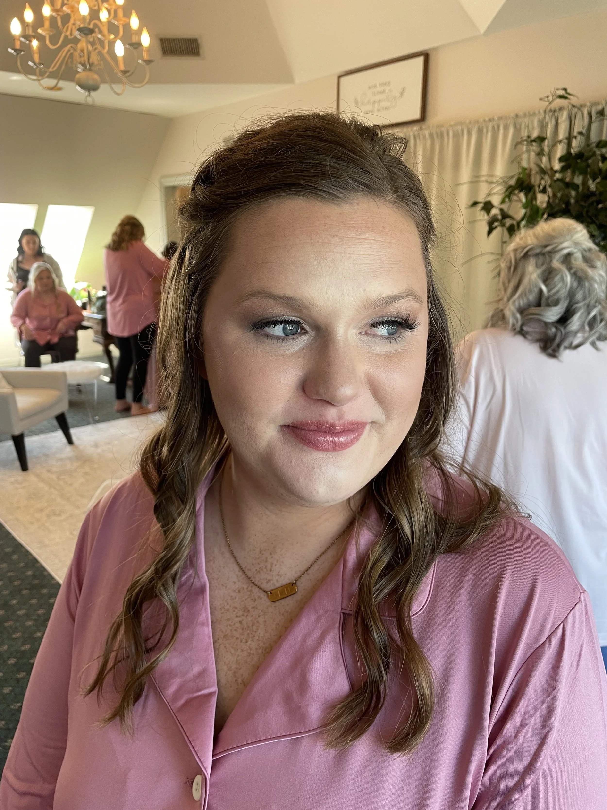 A young woman with brown hair styled in loose waves, wearing a pink satin or silk top and a gold necklace, sitting indoors with a neutral expression. In the background, there are other women, some seated, some standing, and a chandelier hanging from 