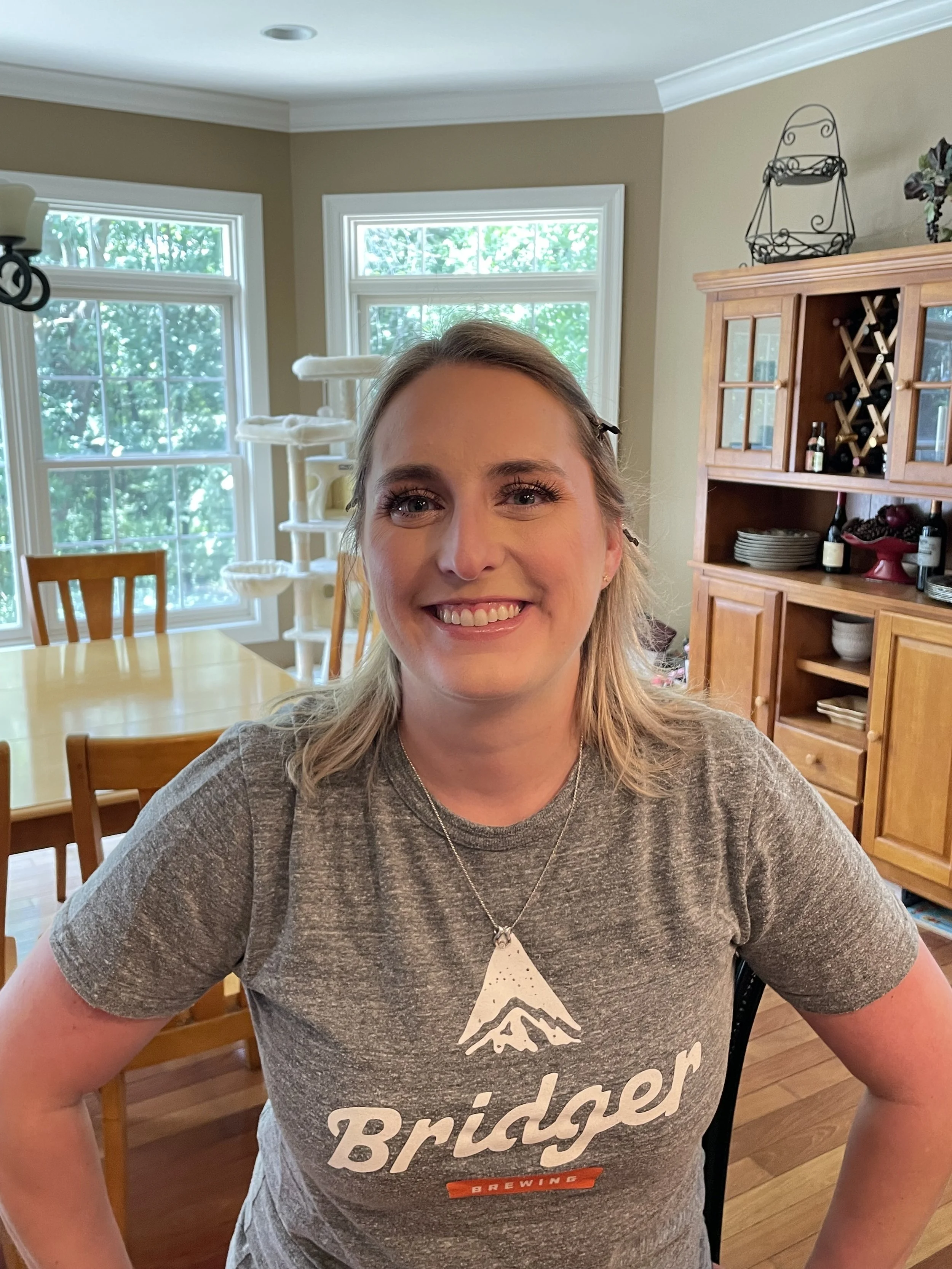 A woman with blonde hair and a gray Bridger Brewing t-shirt sitting at a wooden table in a bright kitchen with large windows and a wooden cabinet in the background.