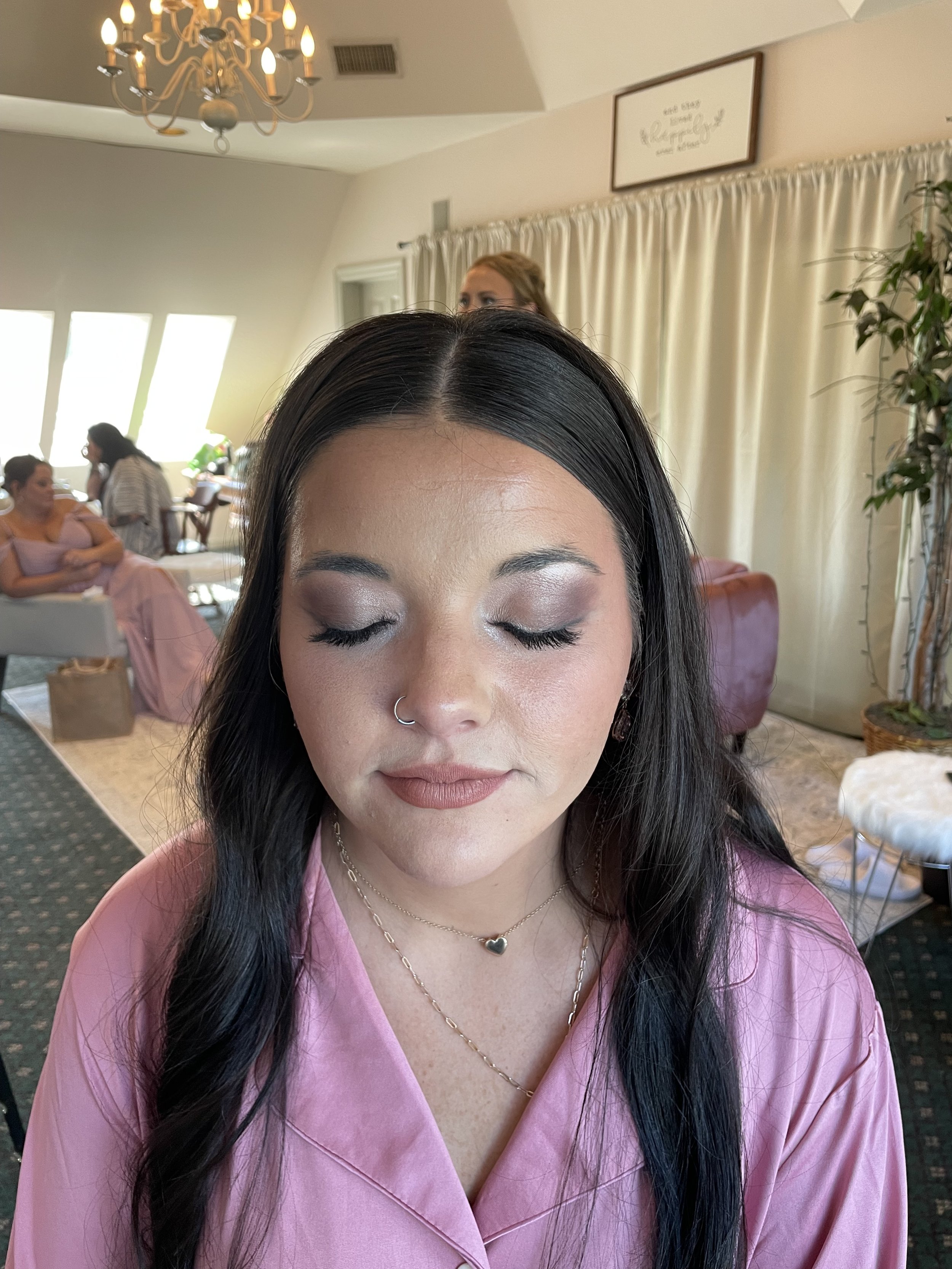 A woman with long dark hair, closed eyes, light makeup, nose ring, wearing pink satin pajamas, a heart-shaped pendant necklace, and earrings, sitting with a calm expression in a well-lit room with other women and a chandelier in the background.