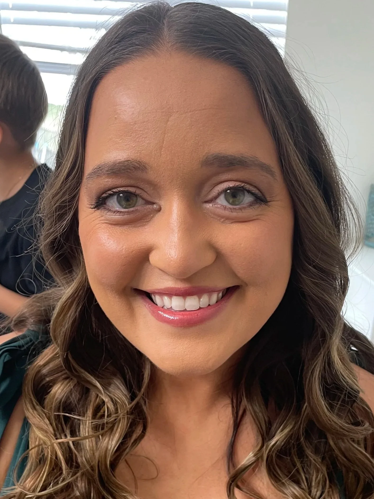 Close-up of a smiling woman with wavy brown hair and light green eyes, in an indoor setting with blinds in the background.