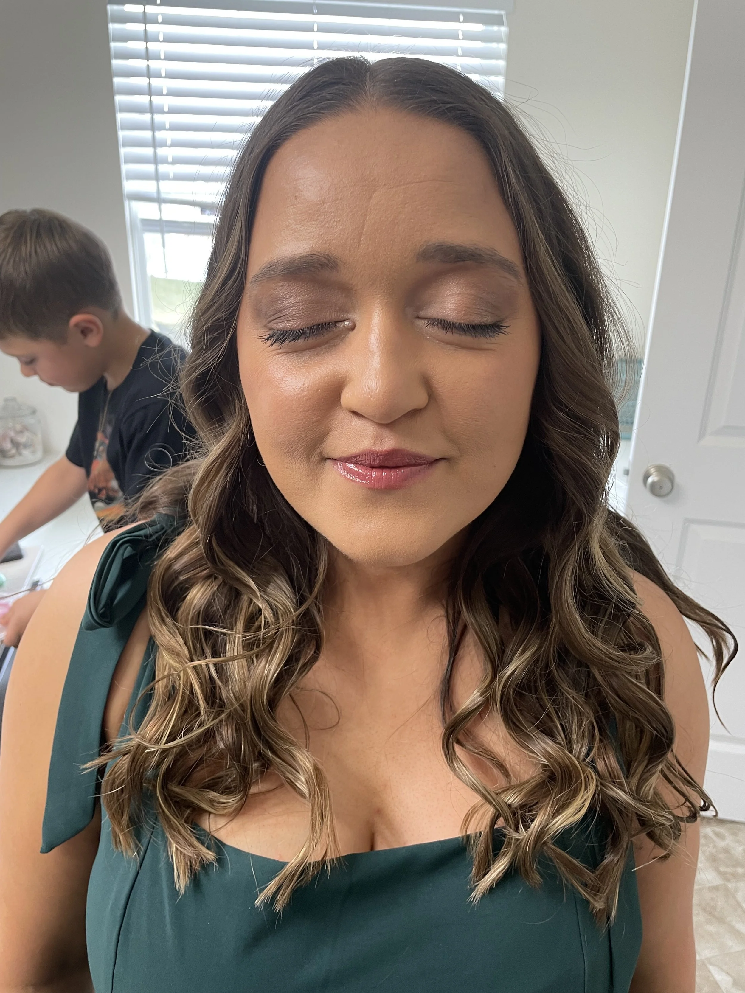 A woman with wavy brown hair and closed eyes, wearing makeup and a green sleeveless top, standing in a room with a window and a boy in the background.