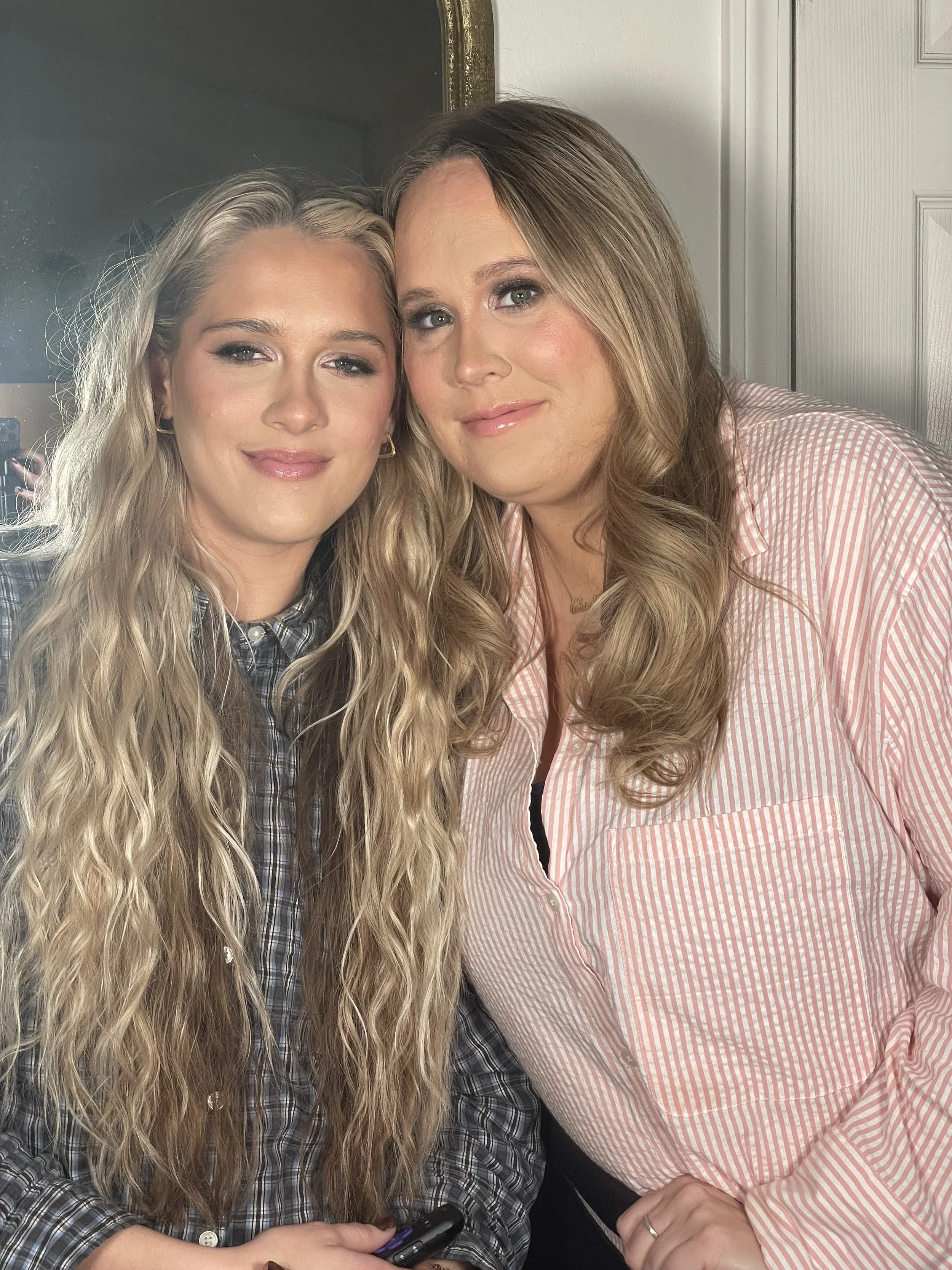 Two women with long, wavy blonde hair smiling and posing together indoors.