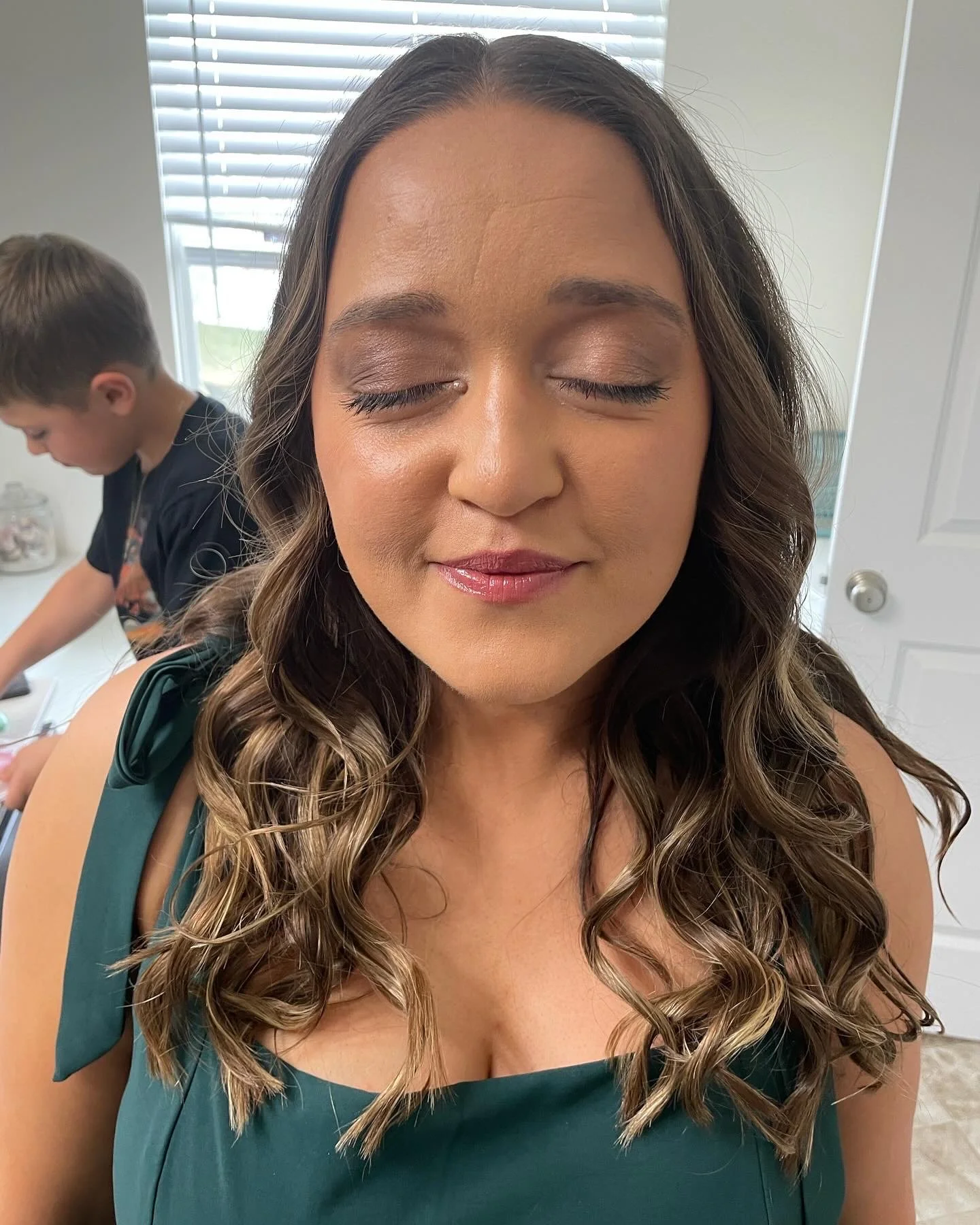 A woman with long wavy hair and makeup, wearing a dark green dress with a bow on the shoulder, closing her eyes and smiling softly. In the background, a young boy is engaged in an activity at a table, near a window with blinds.