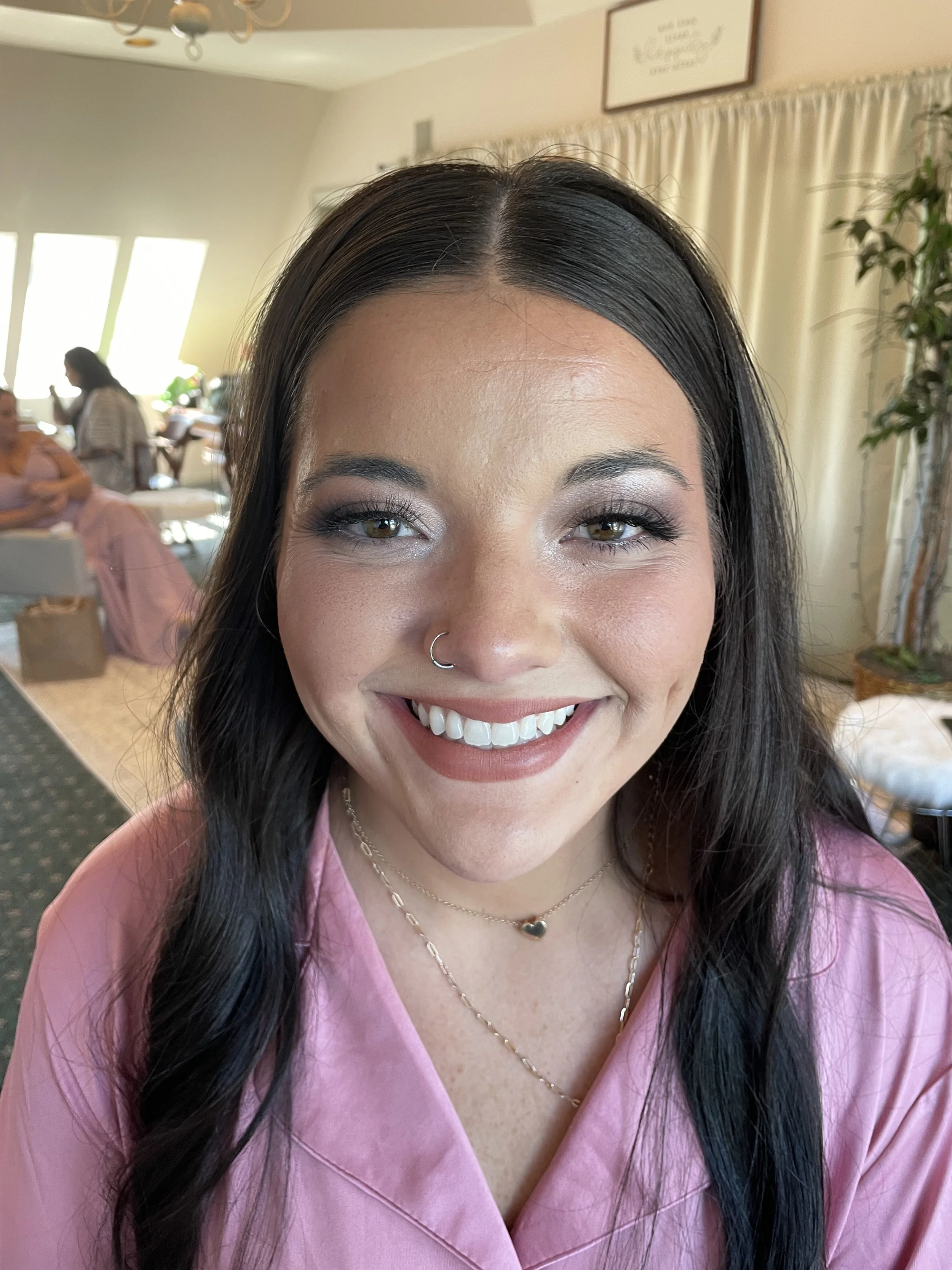 A woman with dark hair, light makeup, and a nose piercing smiling in a room with people in the background.
