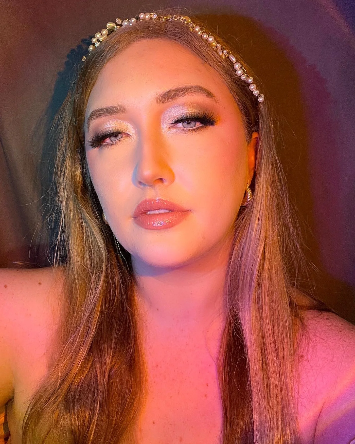 A close-up of a woman with long, light brown hair wearing a pearl headband and earrings, with makeup including eye shadow and glossy lips, winking at the camera.