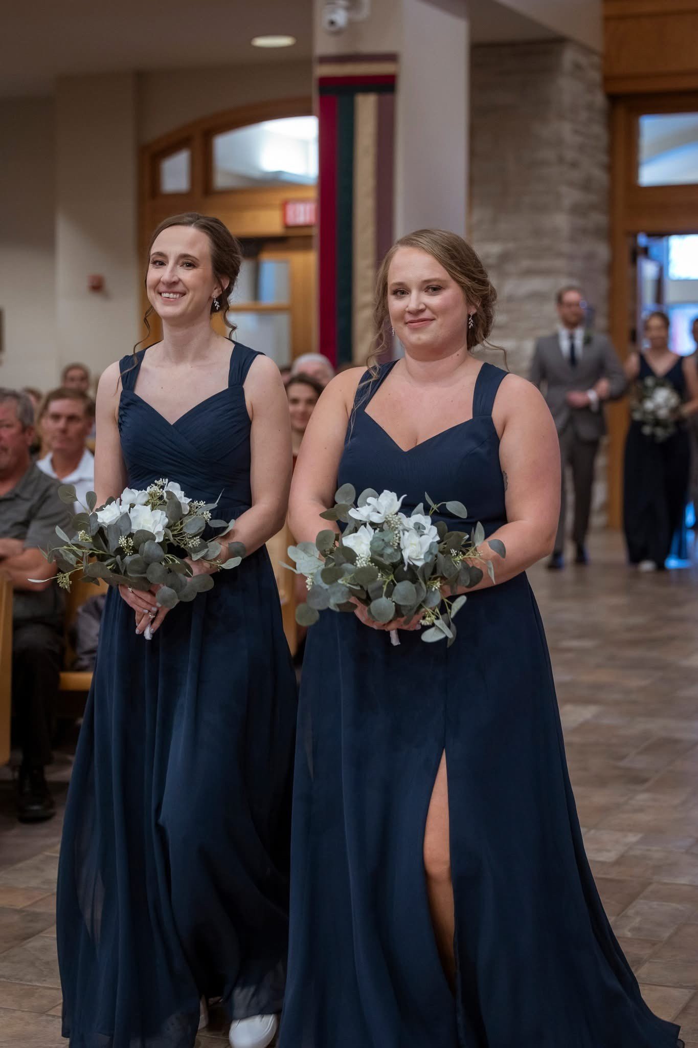 Two women in navy blue bridesmaid dresses walking down the aisle at a wedding, holding bouquets of white flowers and green foliage, with guests seated on either side.
