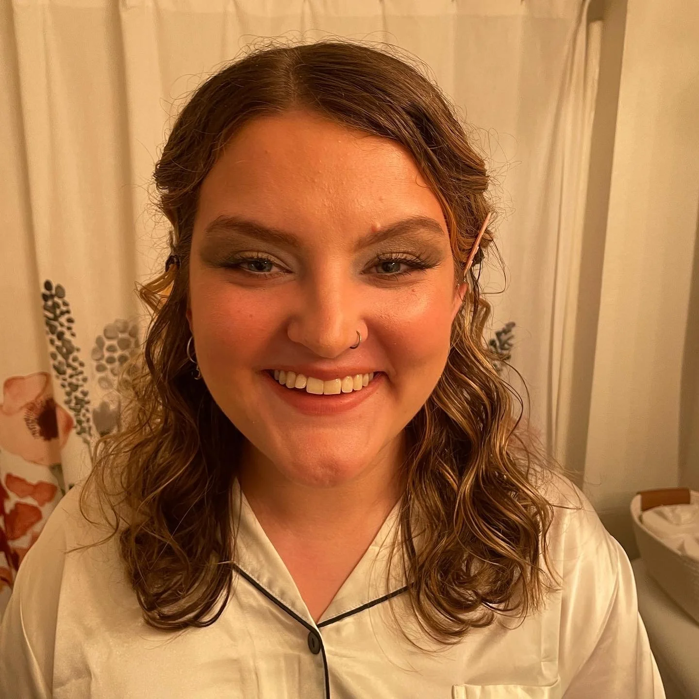 A woman with wavy brown hair, wearing a cream-colored satin pajama top, smiling at the camera with makeup and a nose piercing.