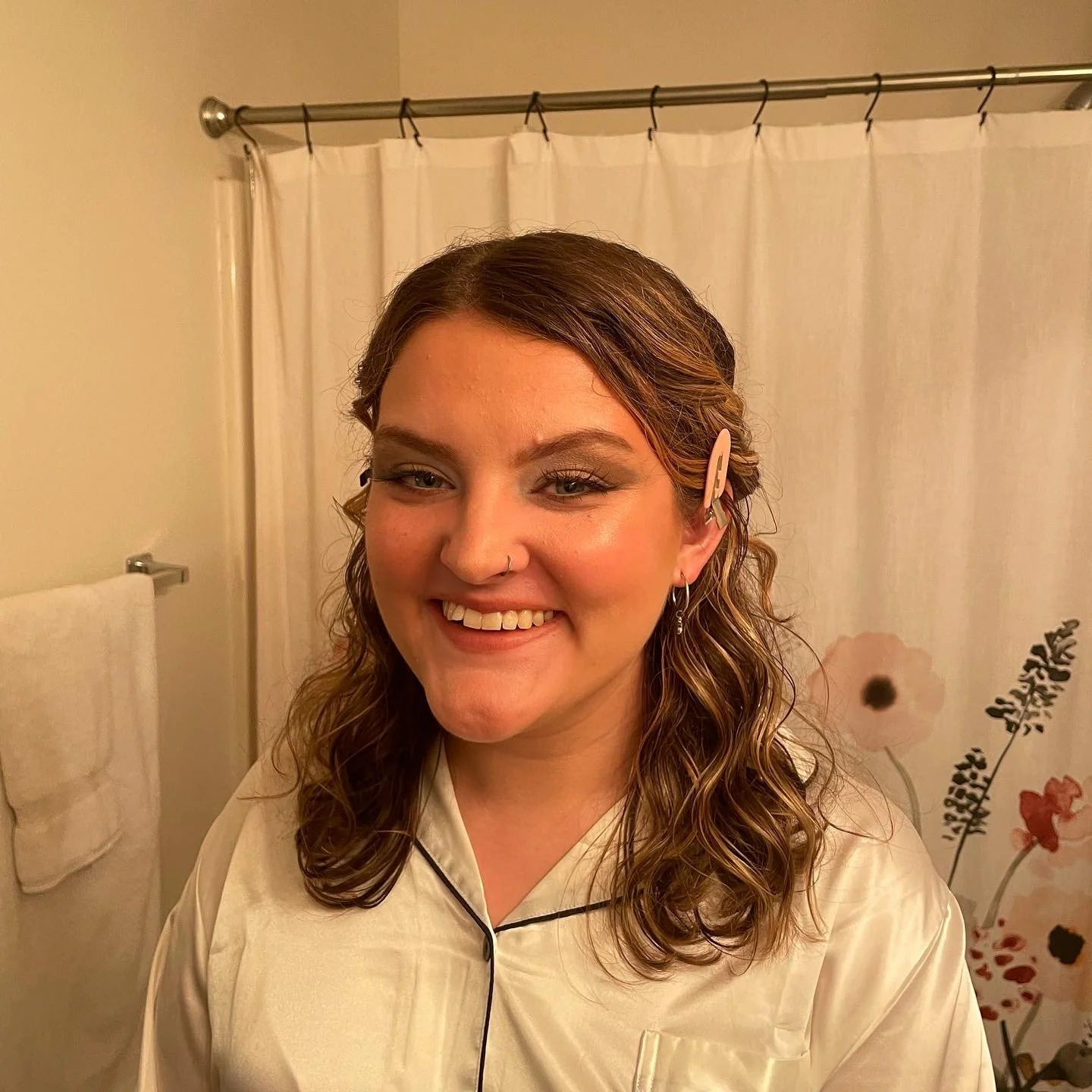 A smiling woman with curly brown hair and a hair clip, standing in a bathroom in front of a shower curtain with floral design, wearing a cream silk pajama top.