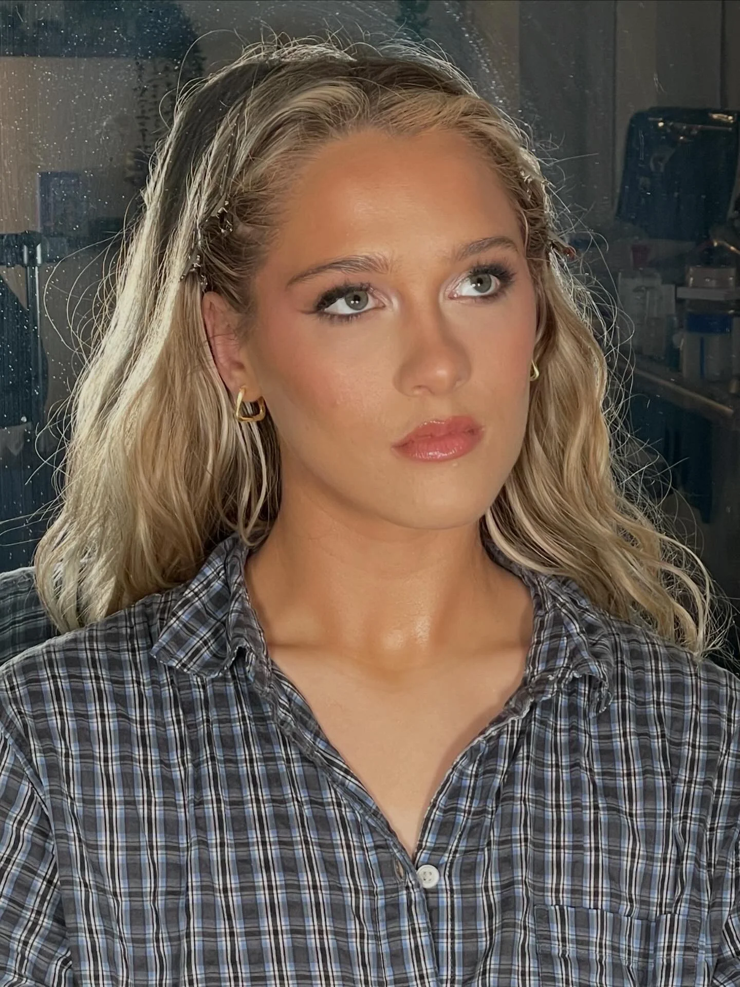 A woman with long curly blonde hair, wearing gold hoop earrings and a blue plaid shirt, looks thoughtfully to the side. The background appears to be a cluttered room with various objects.