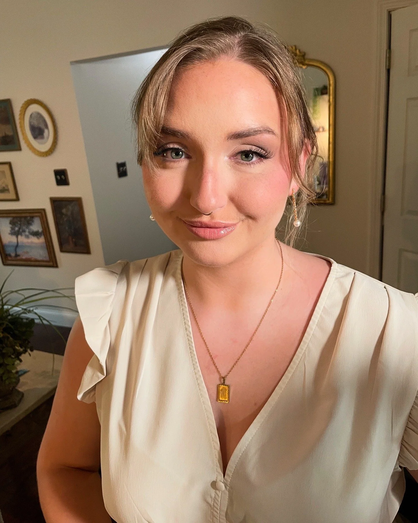 A young woman with light brown hair and green eyes smiling, wearing pearl earrings and a gold necklace, in a room with framed artwork on the walls.