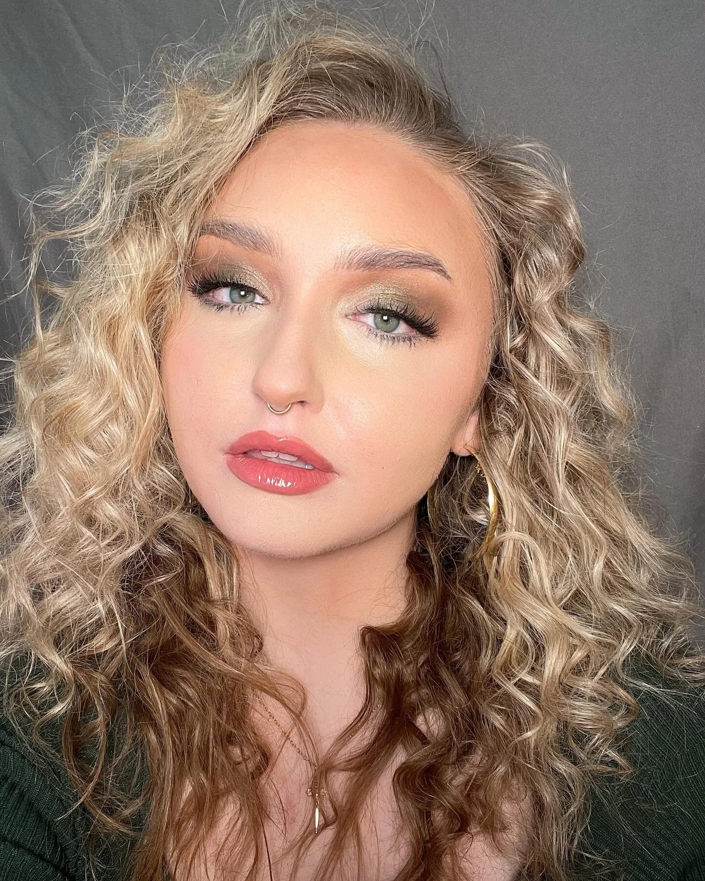 Close-up of a woman with blonde, curly hair, makeup, and a septum ring, wearing gold hoop earrings and a dark top.