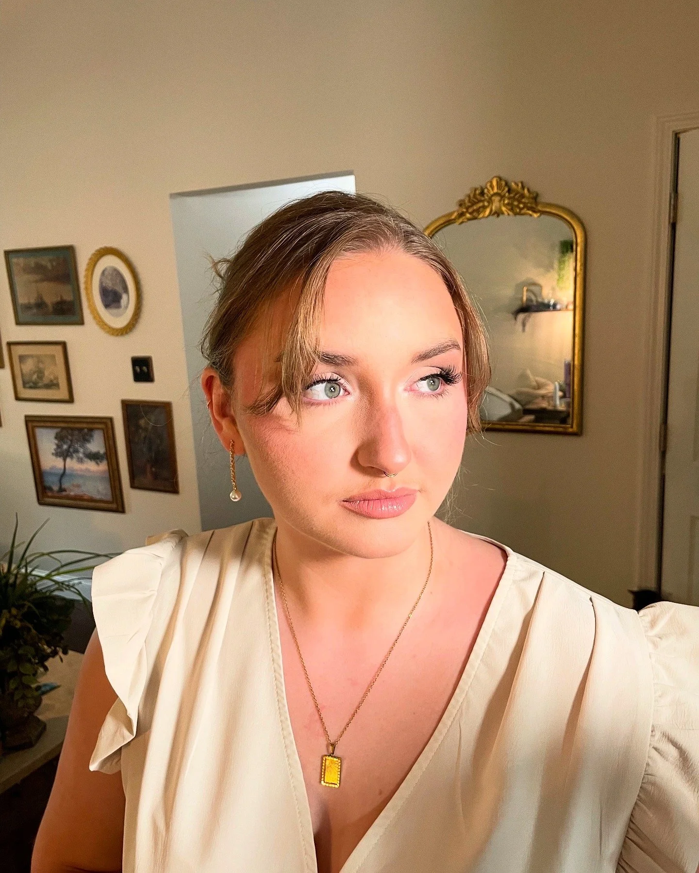 A young woman with light skin, green eyes, and light brown hair styled with a middle part and soft waves. She is wearing a cream-colored top with ruffled sleeves, gold earrings with pearls, and a gold necklace with a rectangular yellow pendant. She i