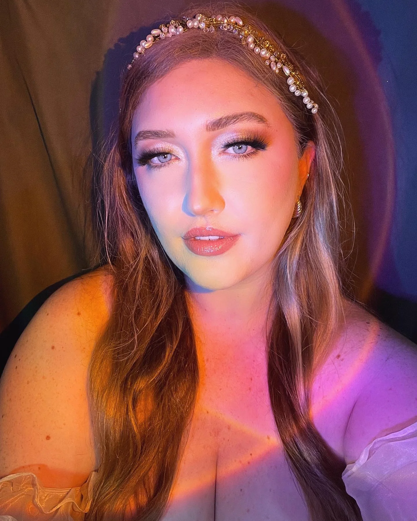 A young woman with long wavy hair, wearing a pearl headband, makeup, and earrings, looking at the camera with a slight smile, illuminated by colorful lighting.