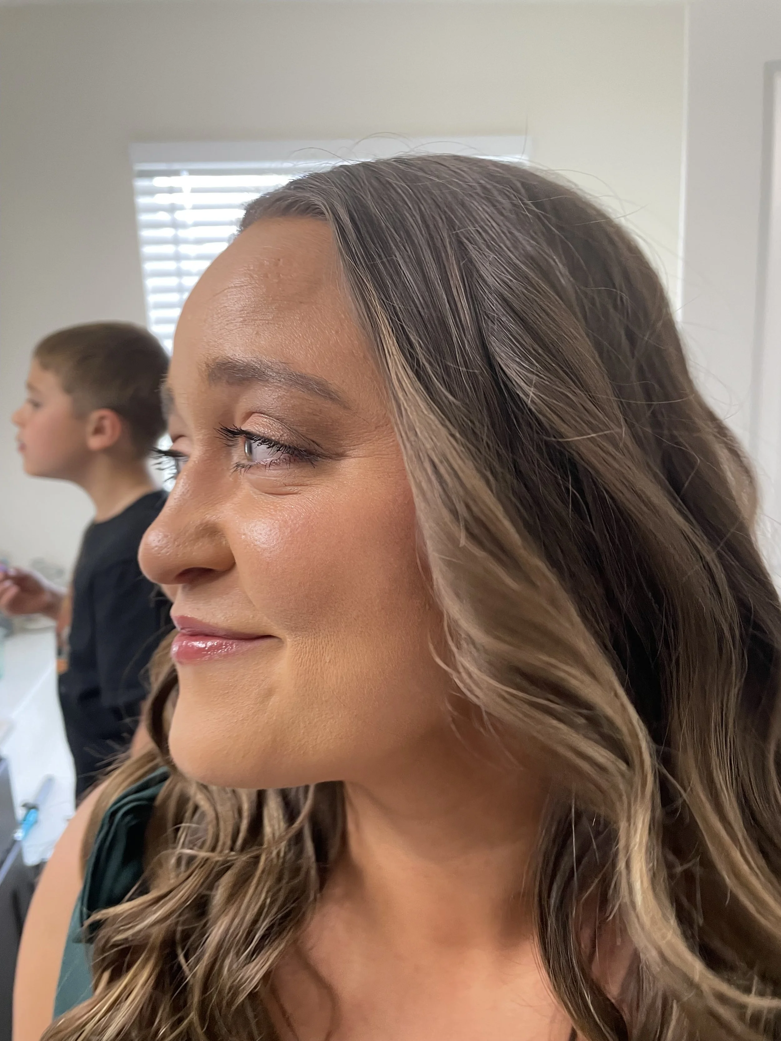 A woman with long wavy hair and makeup, looking to her left. In the background, a young boy with short hair, wearing a black shirt, is blurred and side-profile visible.