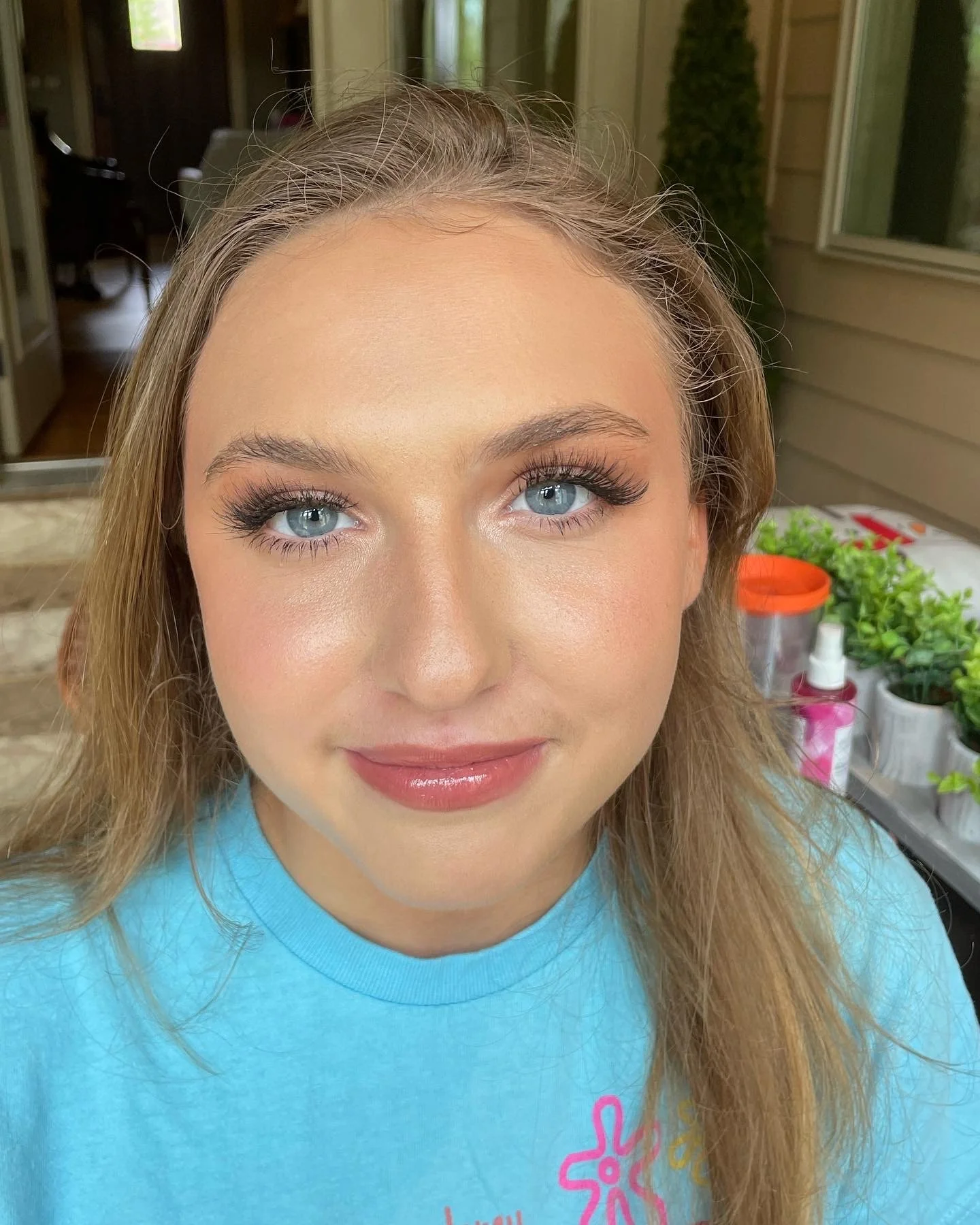 Close-up of a smiling woman with blonde hair and blue eyes, wearing makeup and a light blue shirt, standing outside with plants on a table in the background.