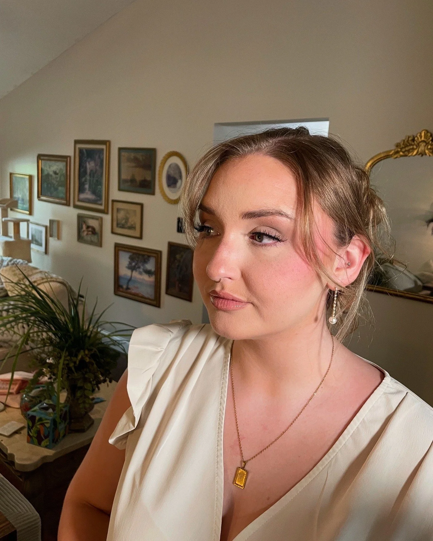 A young woman with light brown hair styled in an updo with loose strands, wearing pearl earrings and a gold necklace with an ornate gold pendant, standing in a room with framed pictures on the wall and a mirror in the background.