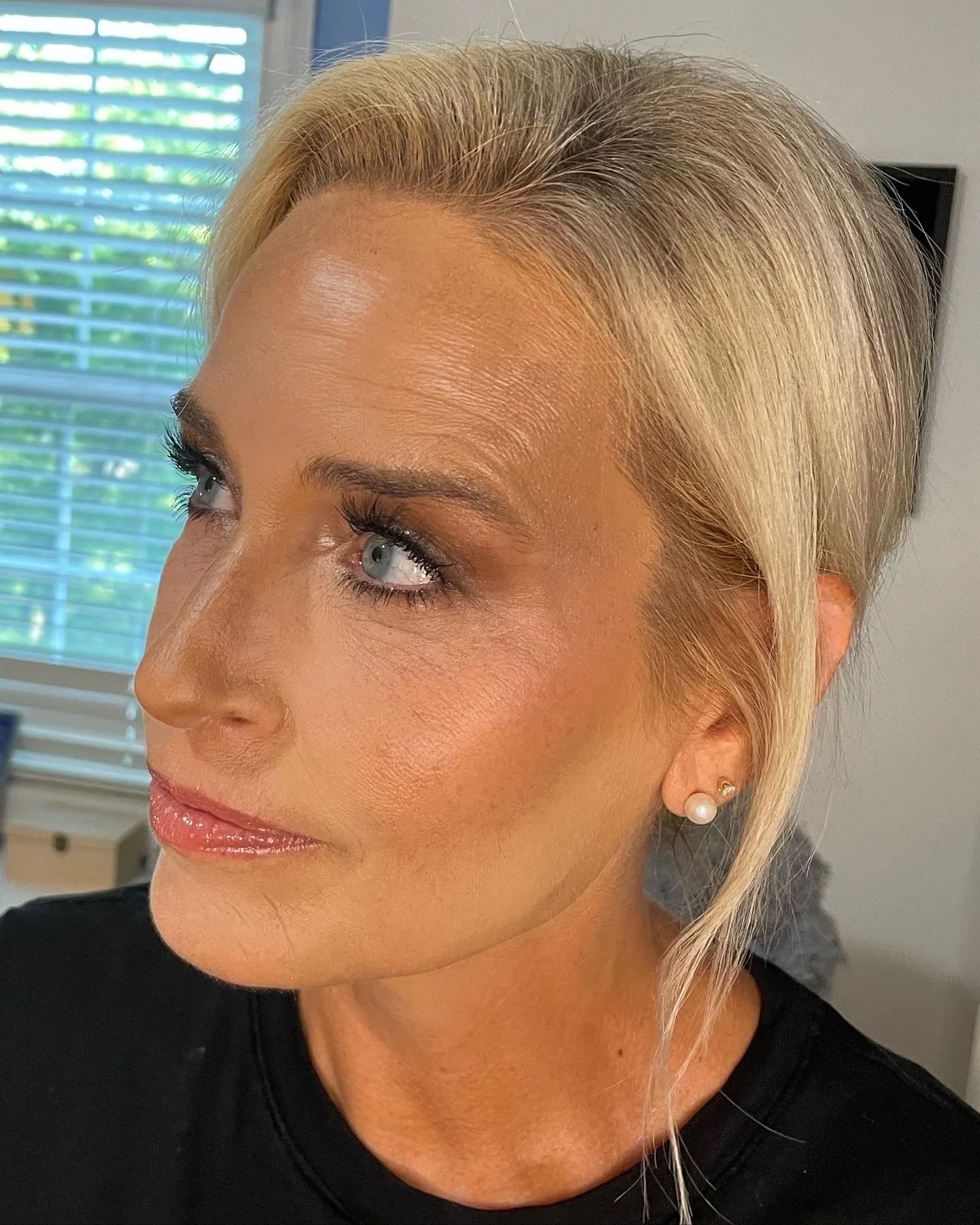 A close-up of a woman with blonde hair styled in a side part, wearing pearl earrings and glossy lipstick, indoors near a window with blinds.
