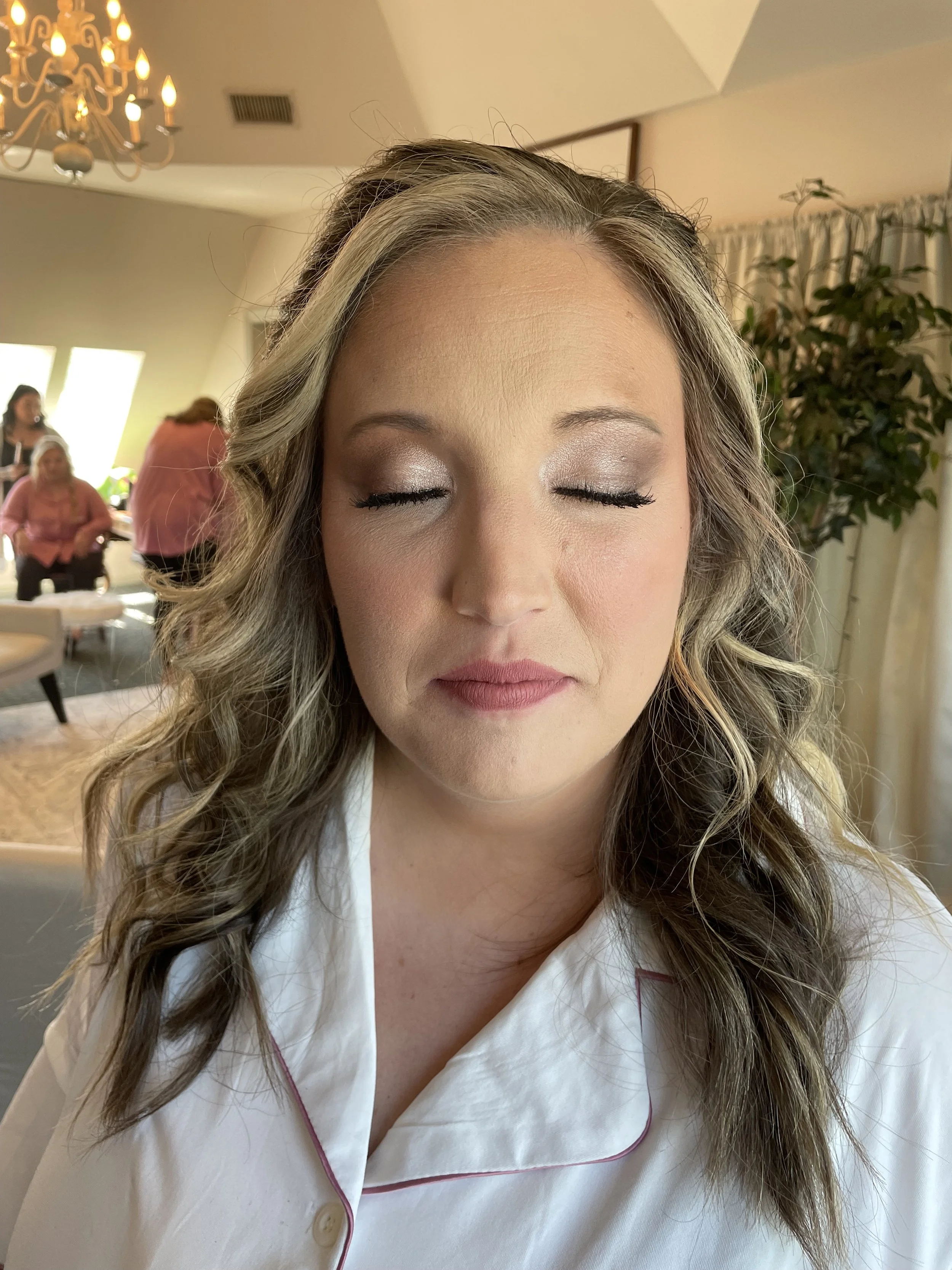Close-up of a woman with her eyes closed, wearing makeup and a white robe with pink piping, in a room with a chandelier, a large window, and other women in the background.