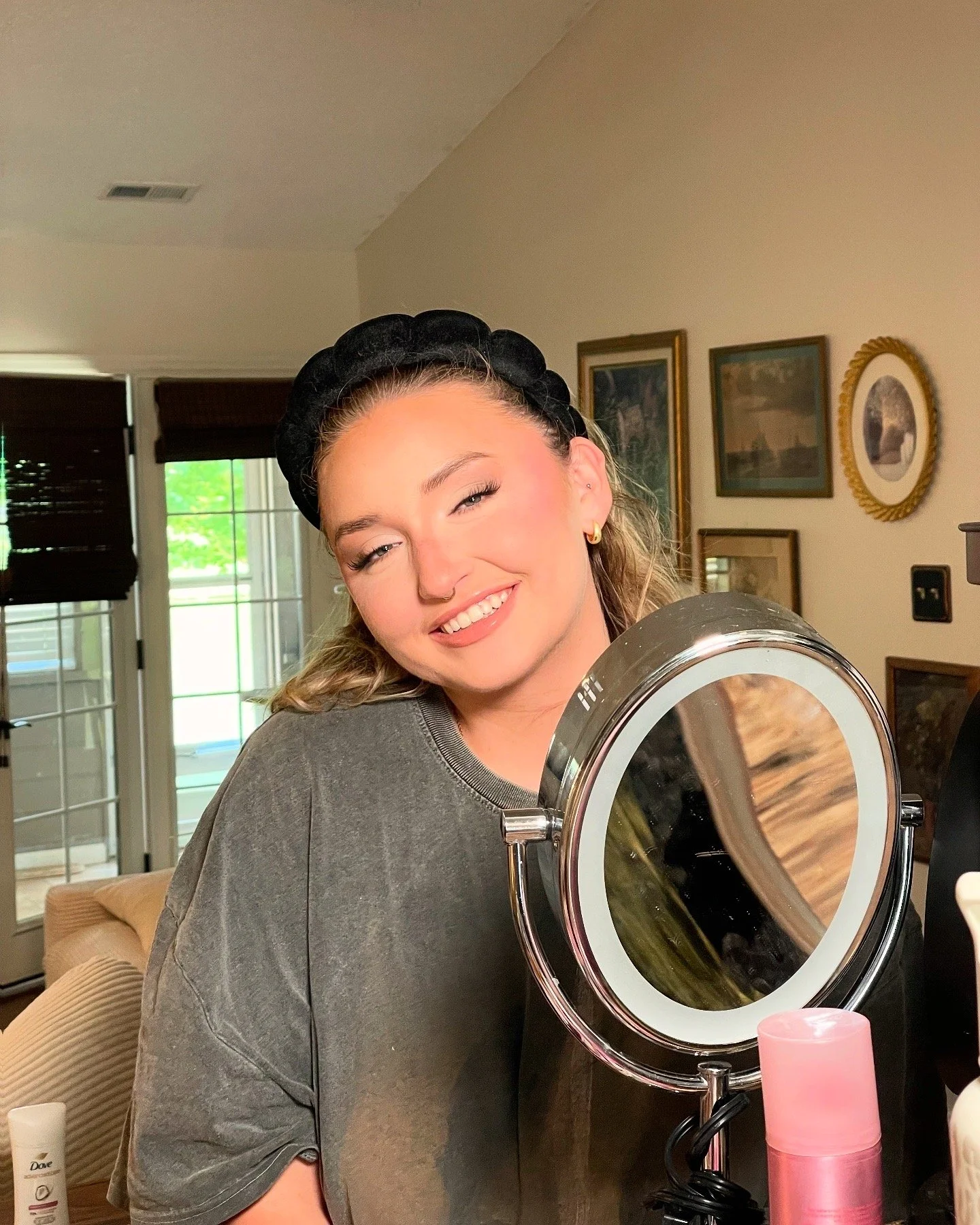 Young woman smiling at a mirror on a vanity table with skincare products, wearing a black headband and gray t-shirt.