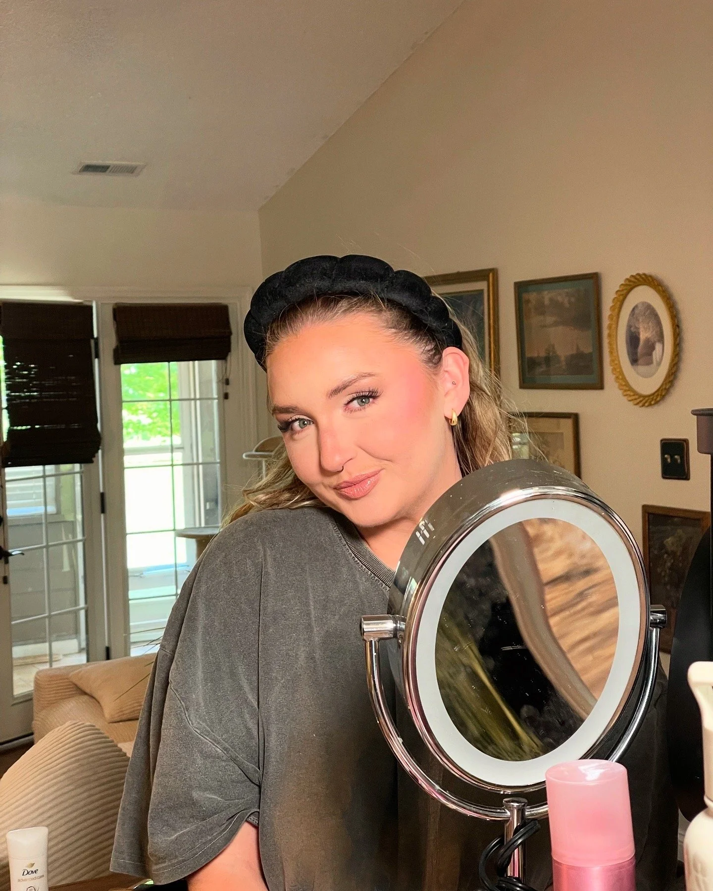 A woman with light makeup and wavy hair, wearing a black headband and a grey shirt, standing in front of a mirror with a content expression on her face.