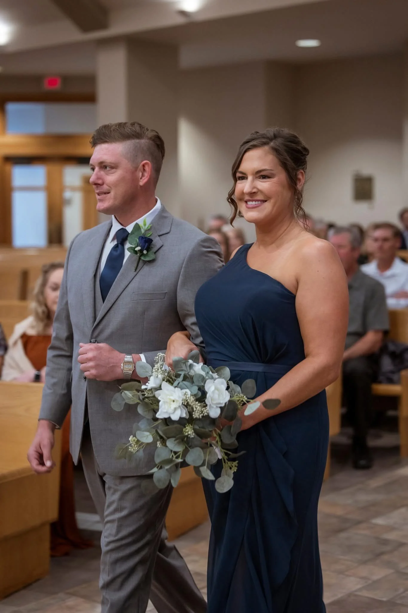 A woman in a navy blue dress holding a bouquet of flowers walking down an aisle during a wedding ceremony with a man in a gray suit and blue tie.