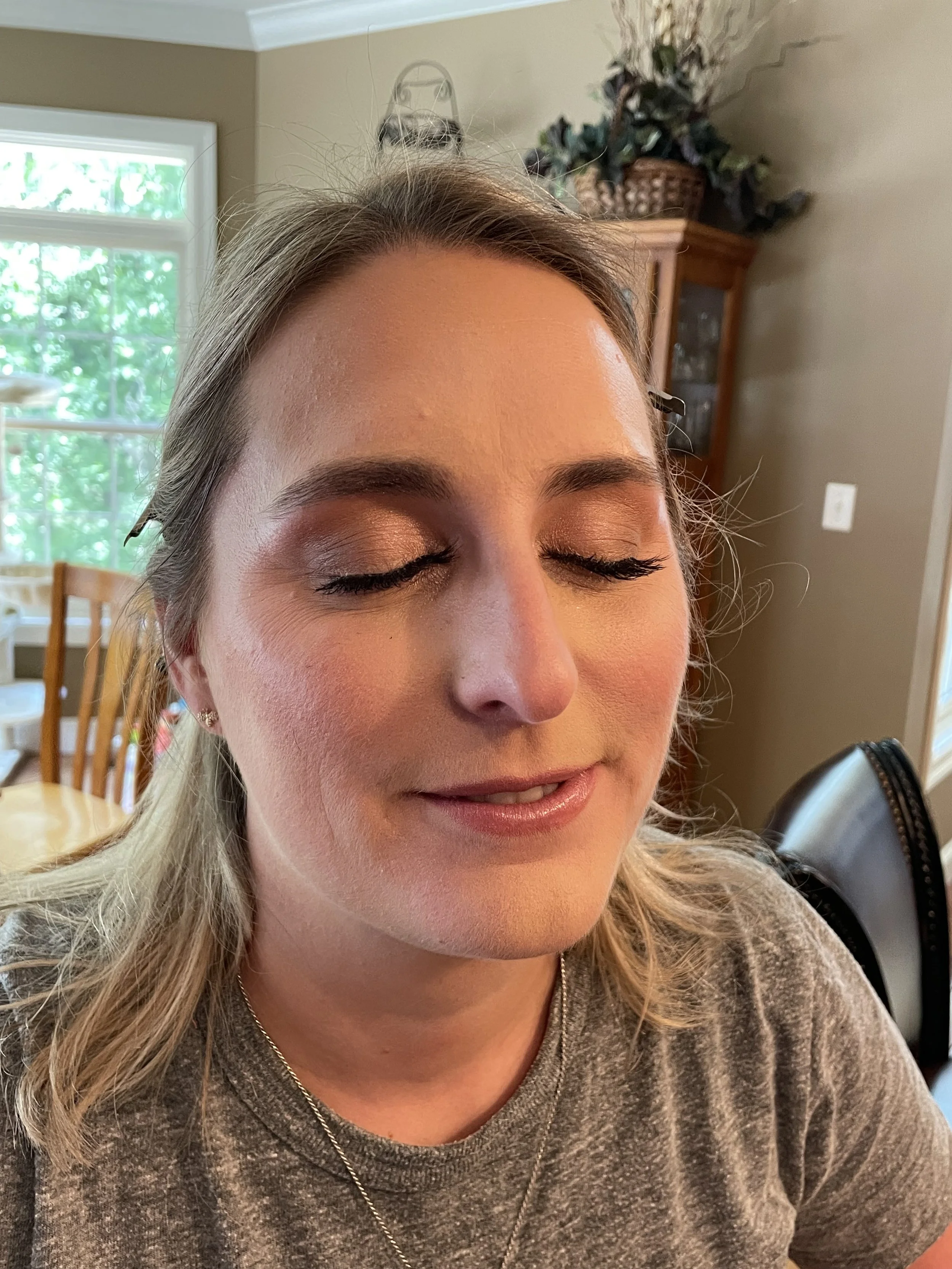 A woman with closed eyes and makeup, smiling gently inside a home with a large window showing greenery outside.